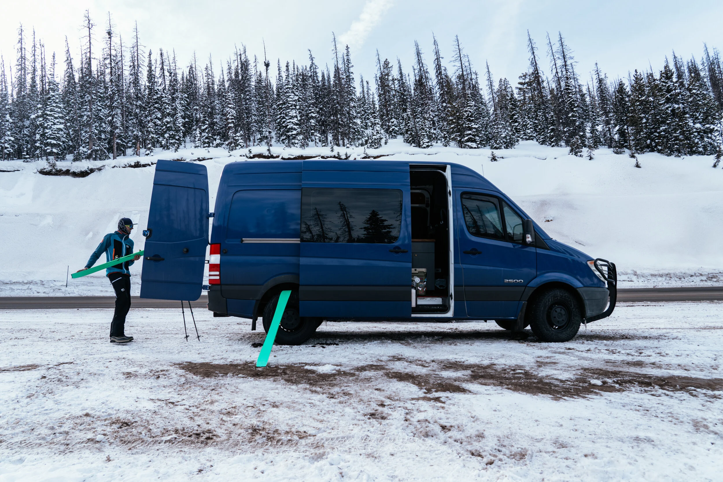 A person standing next to a blue van on a snowy roadside, holding a snowboard, with a snow-covered forest in the background.