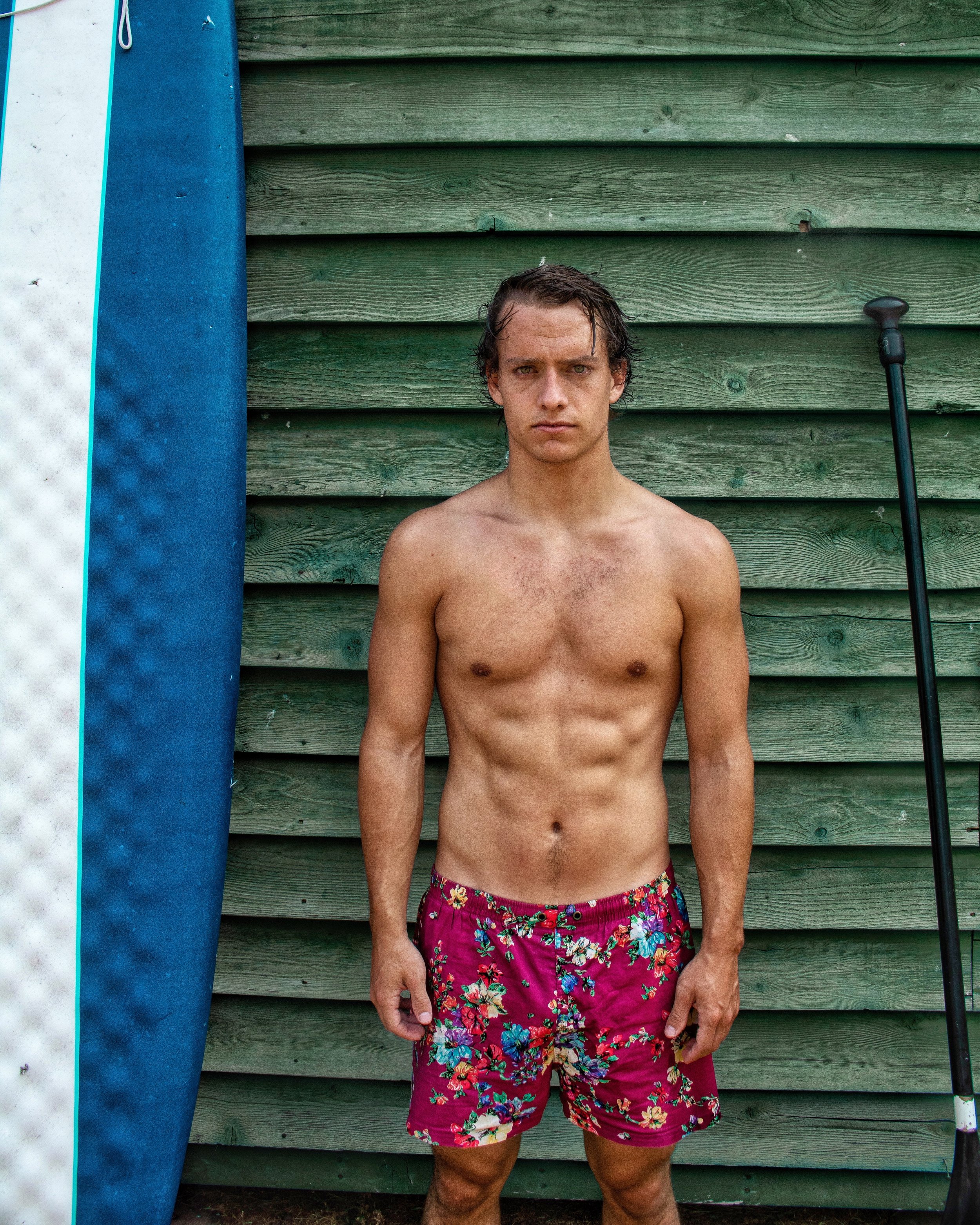 roo standing shirtless next to a paddle board