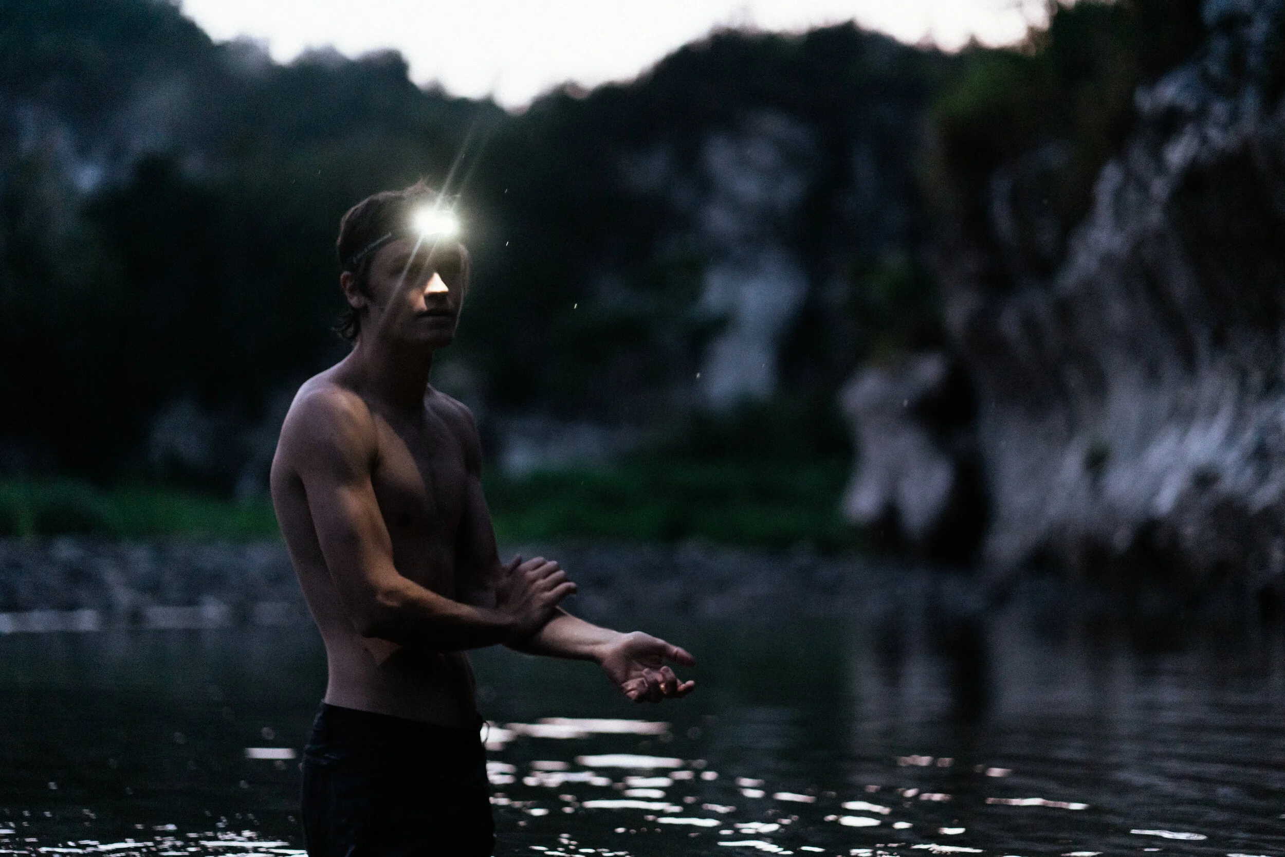 A shirtless man standing in a river during dusk or dawn, with a headlamp on his forehead, surrounded by rocky terrain and trees.