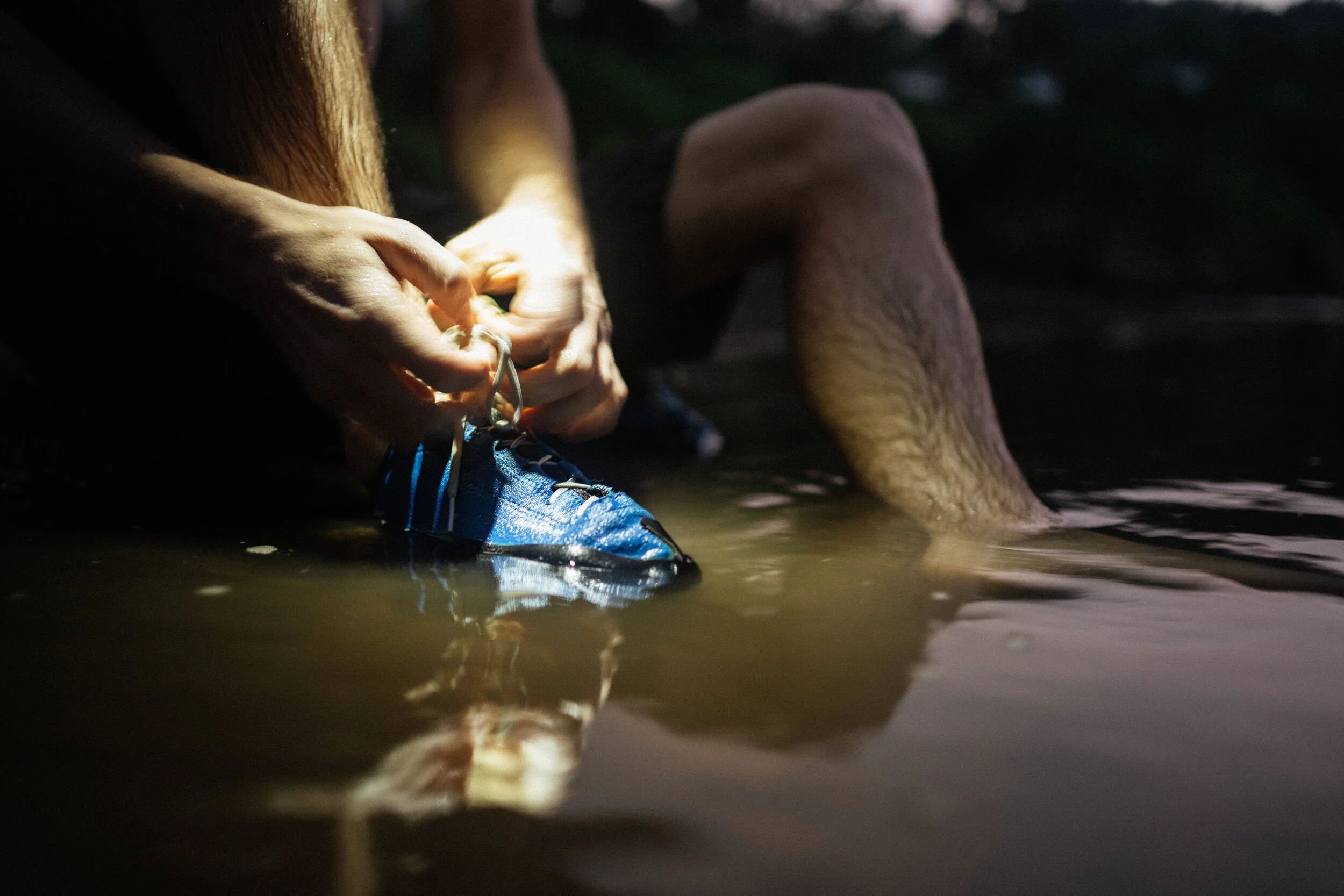 A person is sitting at the edge of a body of water, tying the shoelaces of their blue hiking shoes while their legs are submerged in the water.