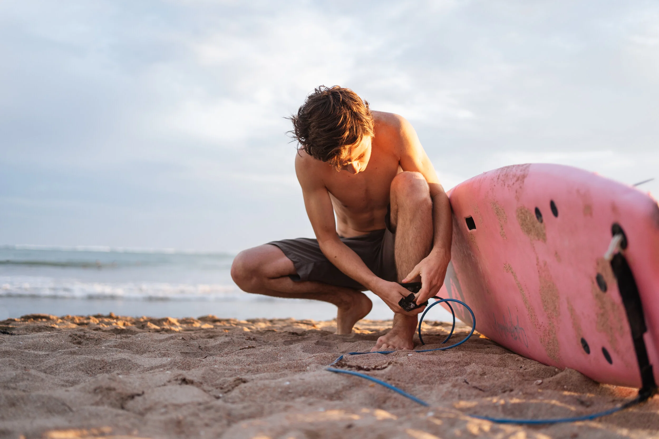 A young man with brown hair, shirtless, crouching on a sandy beach, working on repairing a pink surfboard with some sand on it, with the ocean in the background and a cloudy sky.