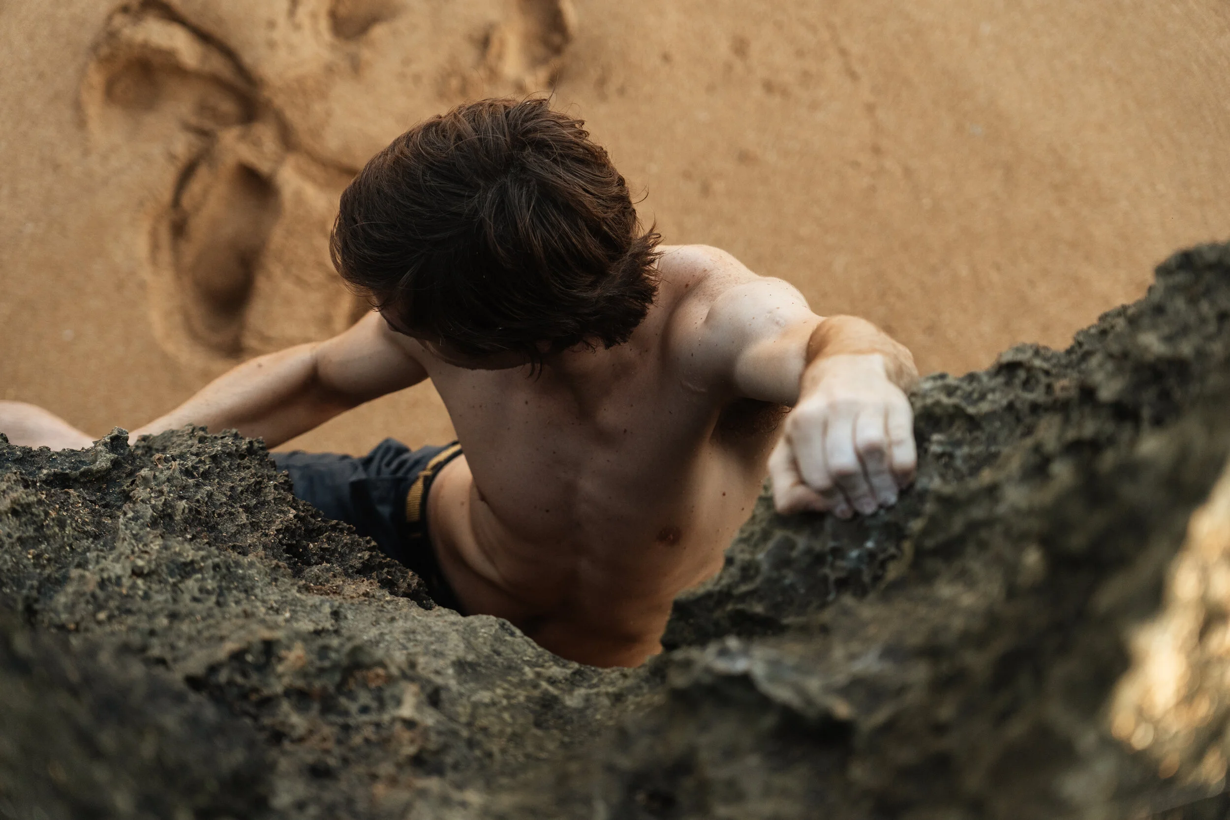 A shirtless man with brown hair climbing a rocky indoor wall.