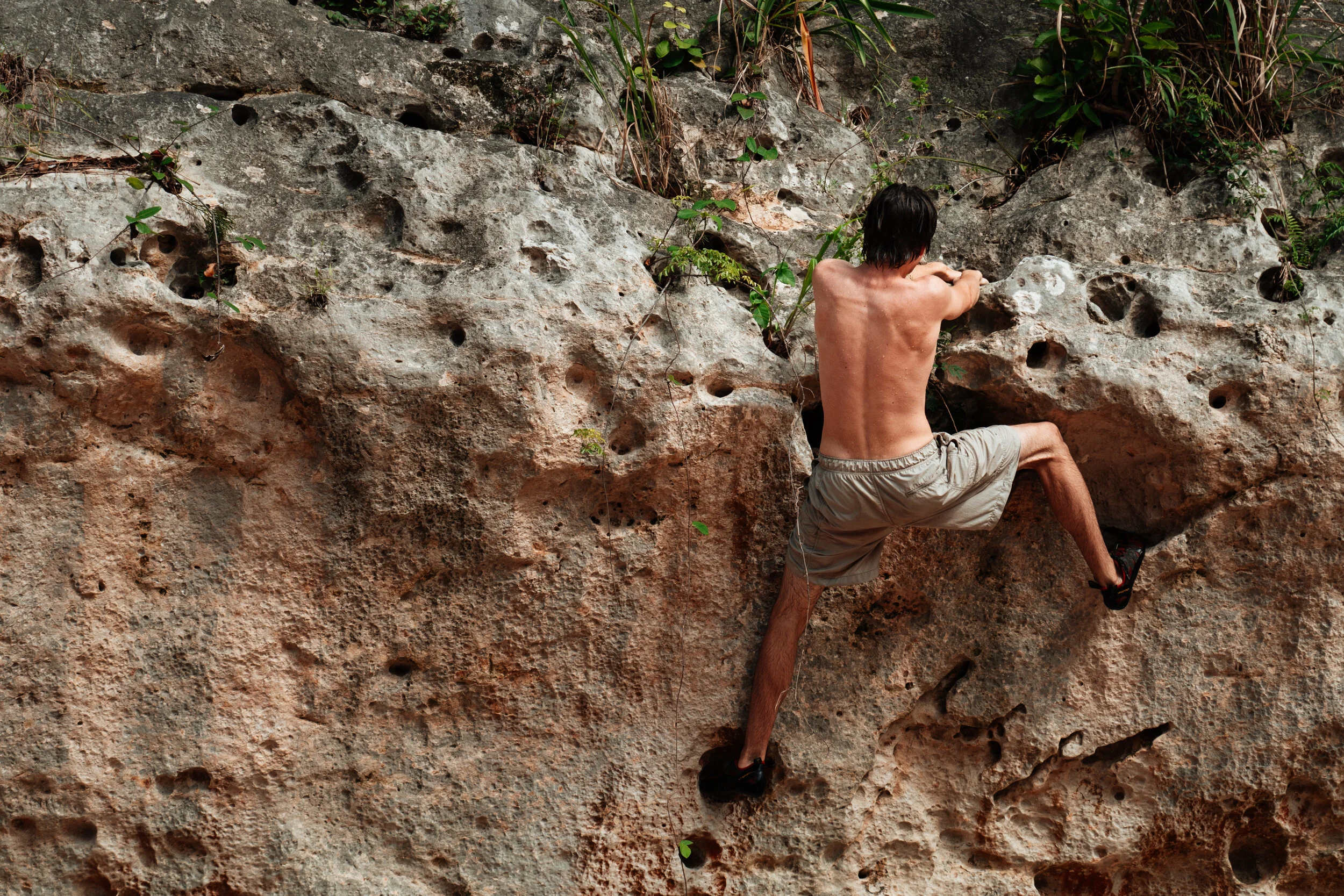 A man with dark hair, wearing beige shorts, is rock climbing barefoot on a textured rock wall outdoors, surrounded by some greenery.