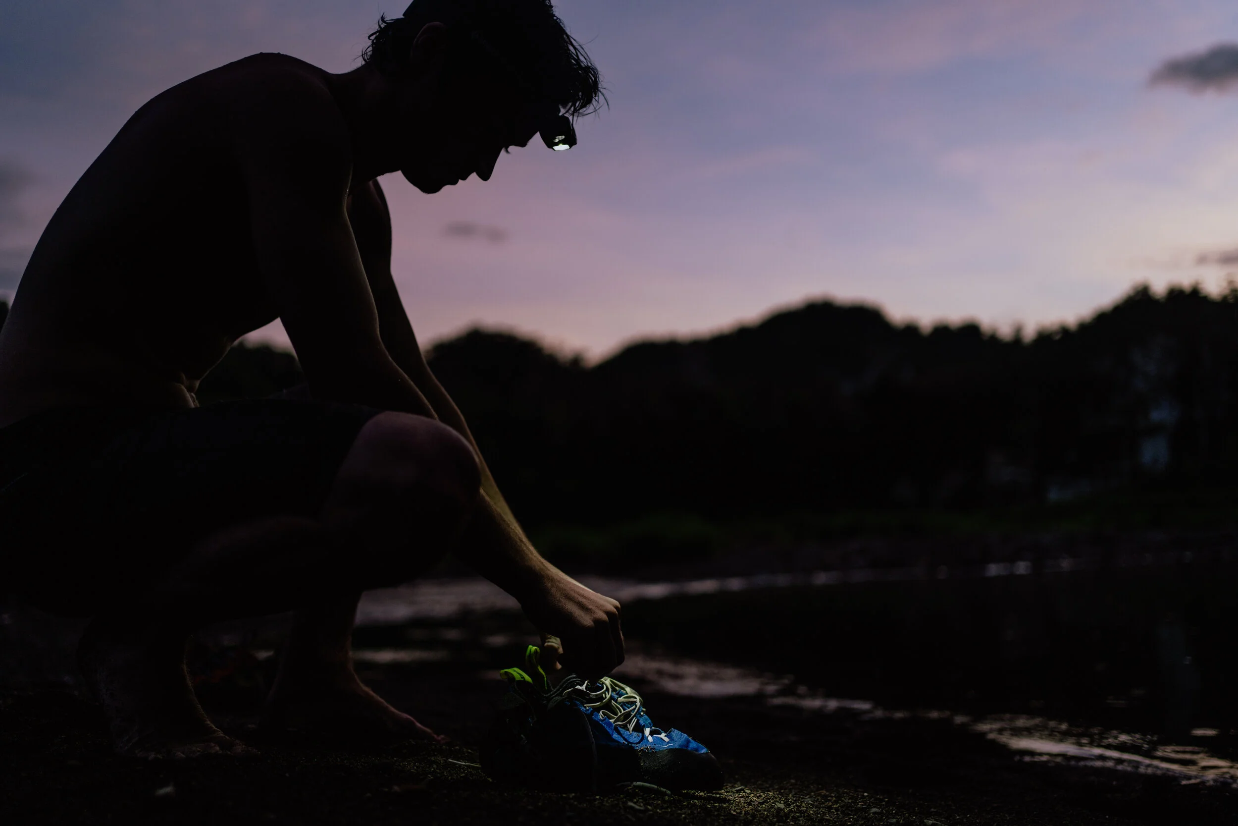 A person squatting outdoors at sunset, tying their running shoes on a dark beach or shoreline.