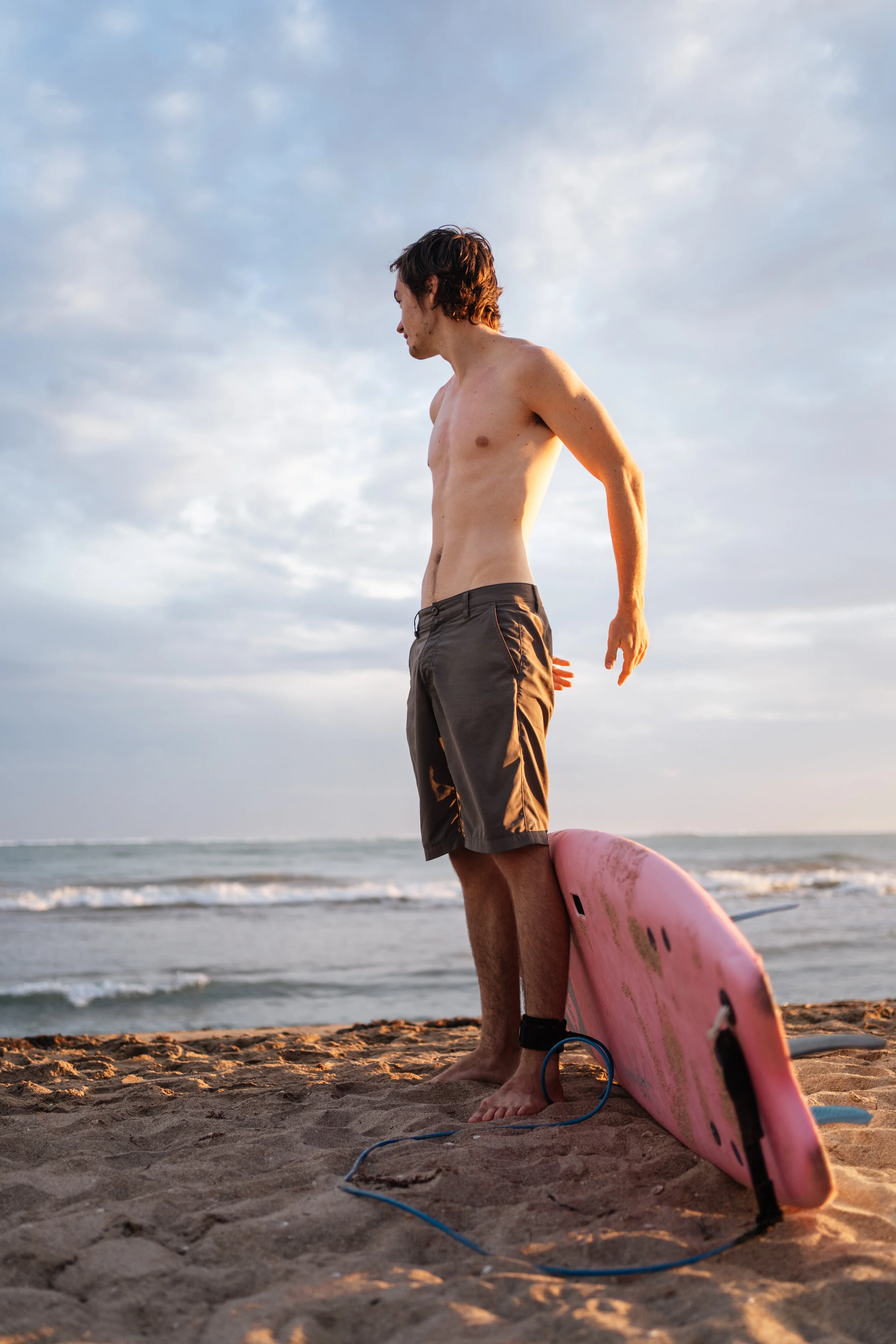 A shirtless man with brown hair standing on a sandy beach next to a pink surfboard, looking down at the ground, with the ocean and a cloudy sky in the background during sunset.