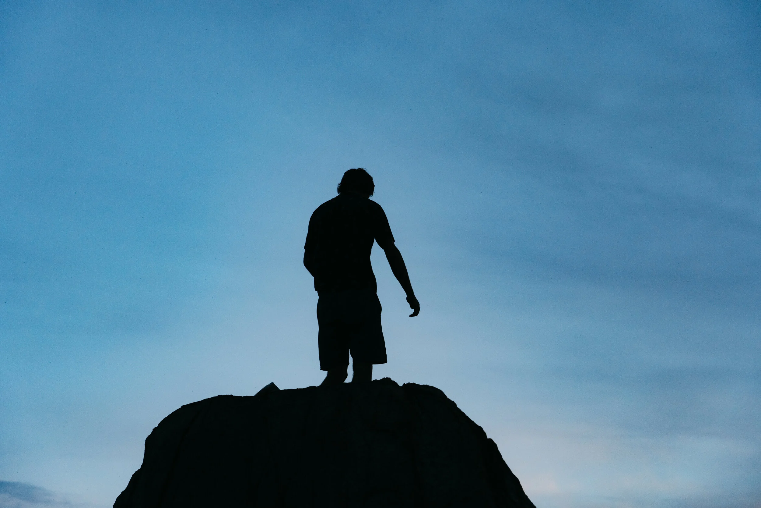 Silhouette of a person standing on top of a rock during dusk or dawn with a blue sky in the background.