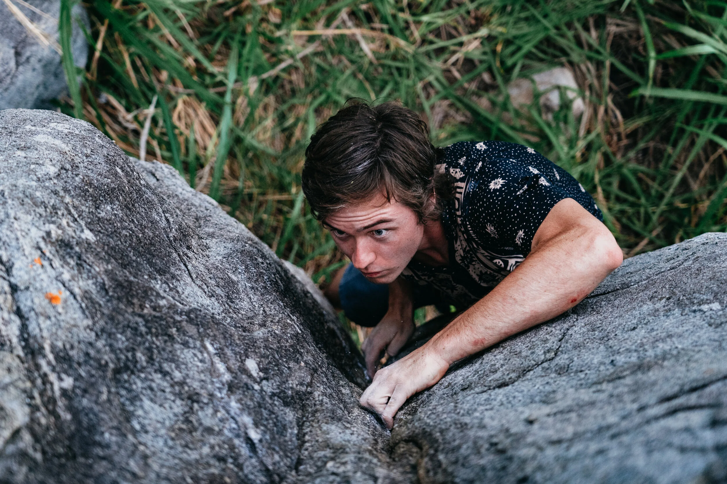 A young man with brown hair climbing a rock face outdoors, surrounded by grass and rocks.