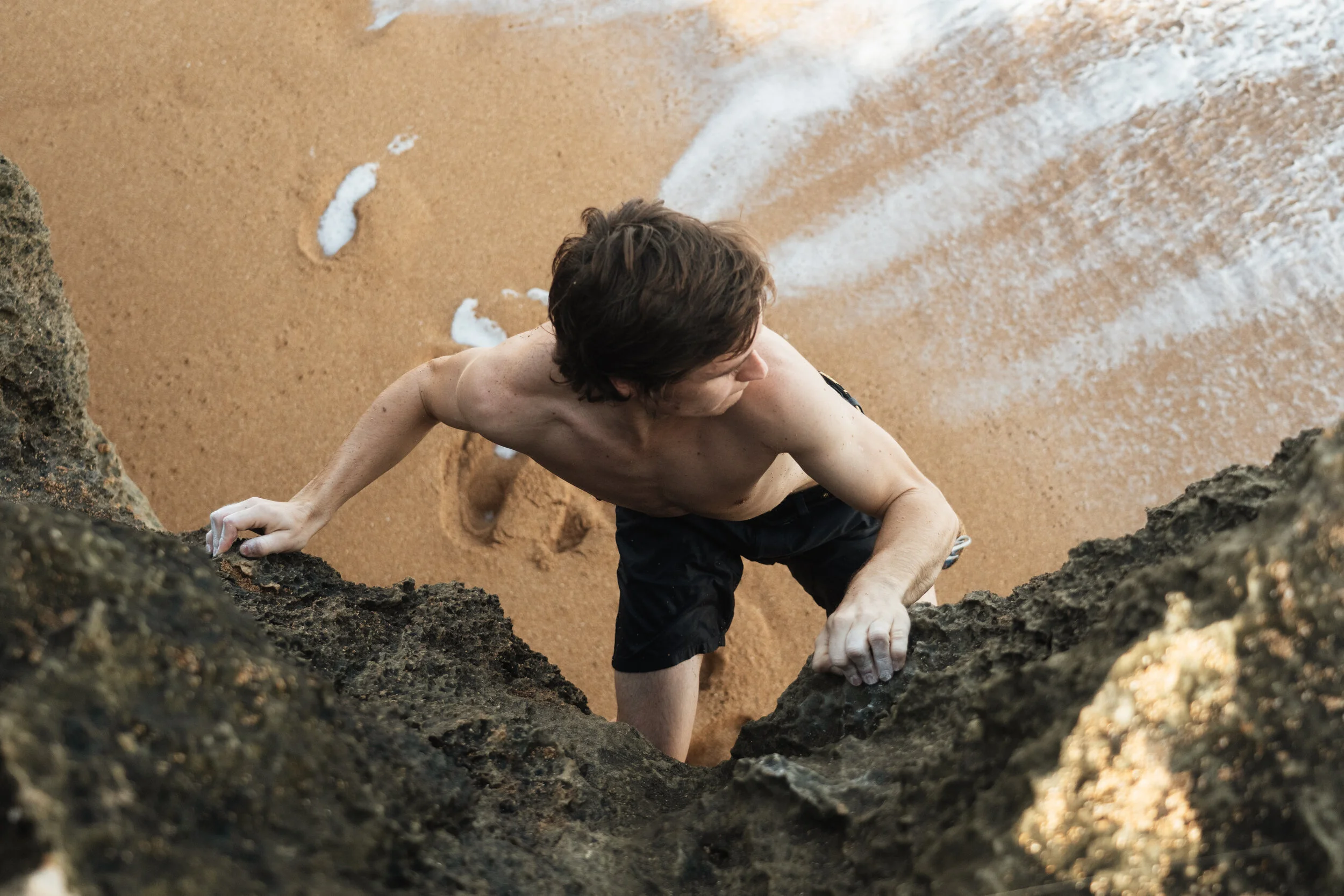 A shirtless man climbing a rocky cliff near the beach, with sand and foam from the ocean visible below.