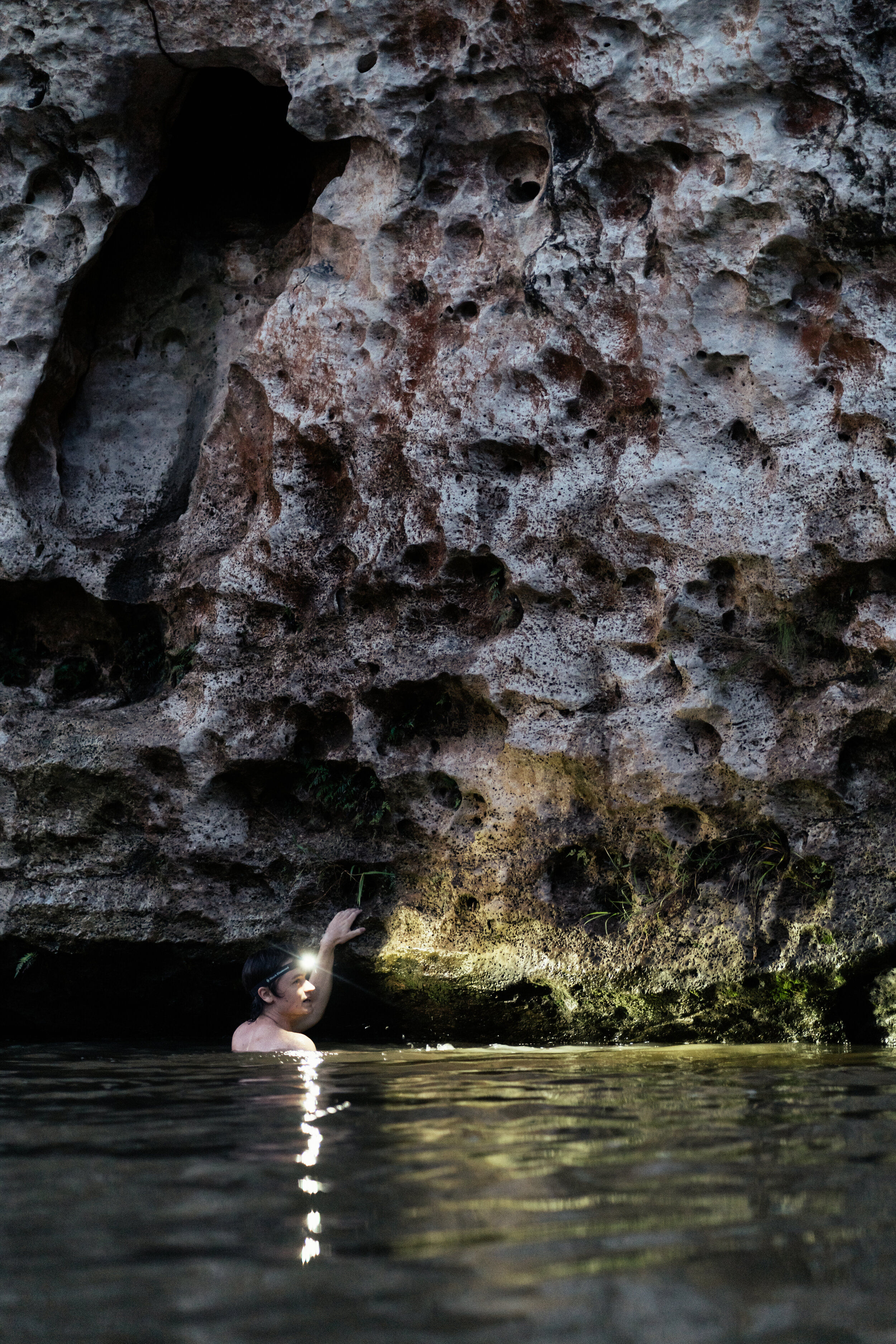 A person swimming in water near a large rock formation with a rough, porous surface and small patches of moss.