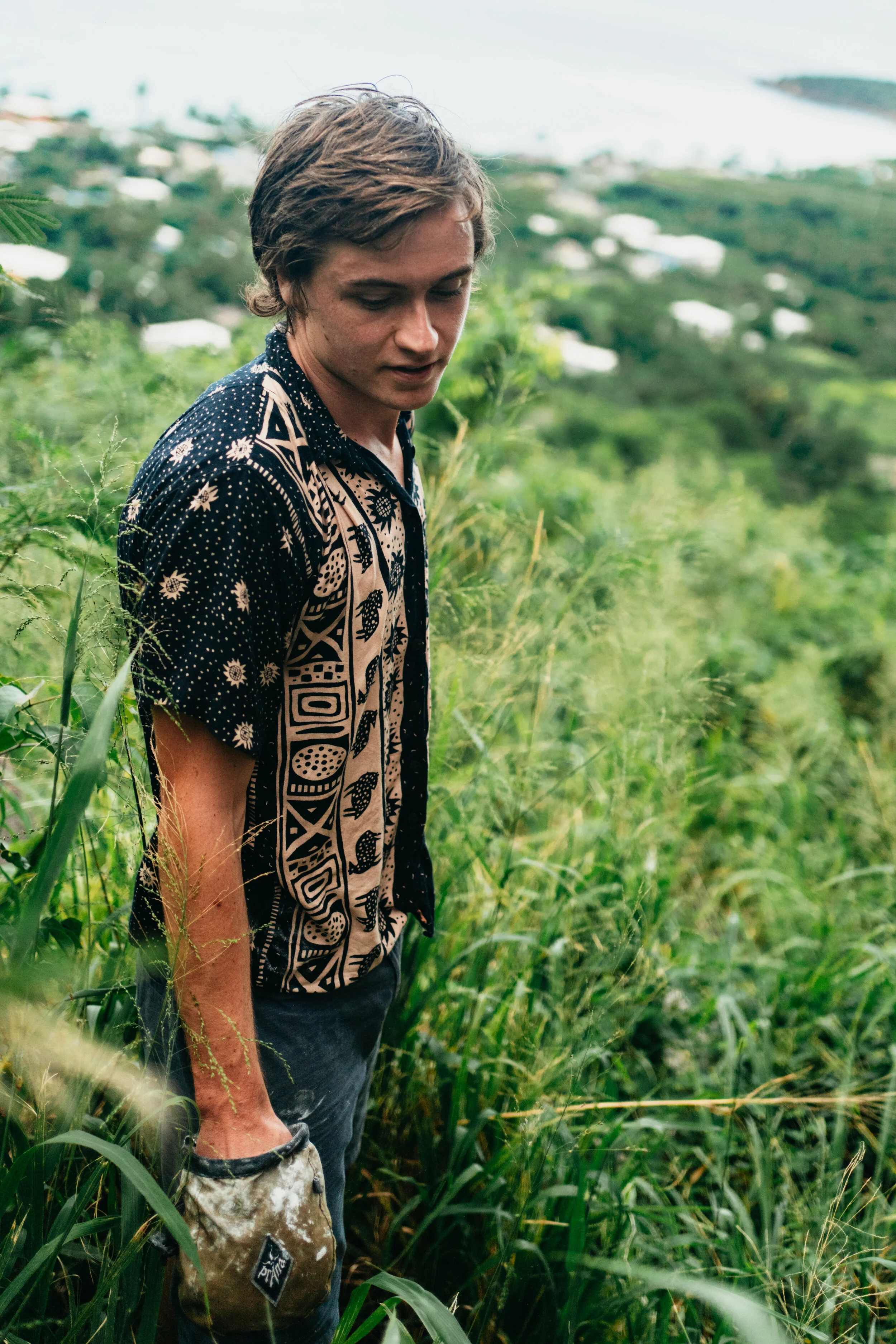 A young man with curly brown hair in a patterned short-sleeve shirt standing in tall green grass with a landscape and water in the background.