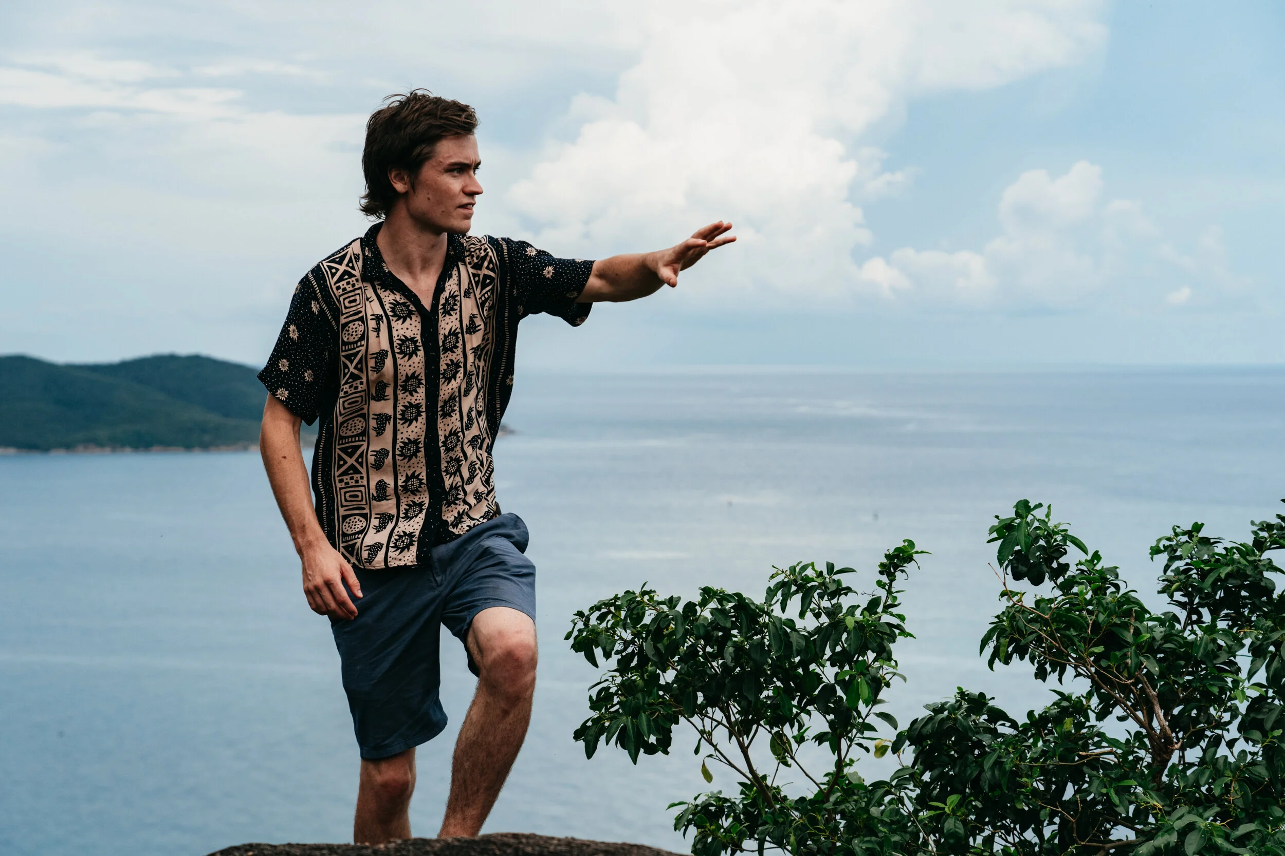 A young man with dark hair, wearing a patterned shirt and blue shorts, standing on a rocky ledge near the ocean, reaching out with his right arm with a cloudy sky overhead.
