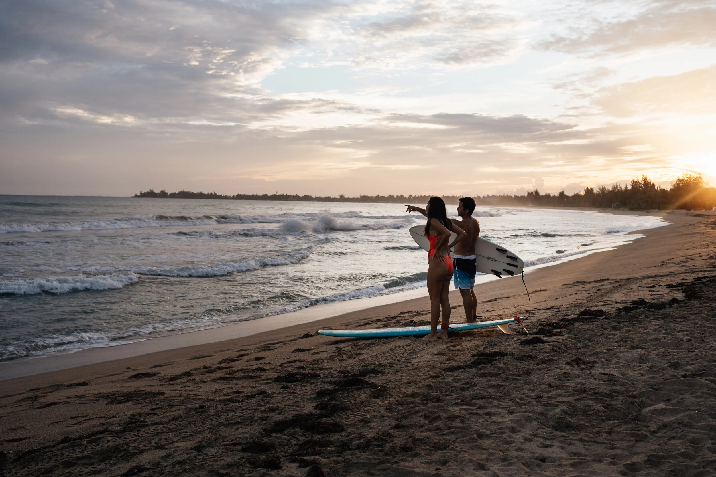 A man and woman standing on the beach holding surfboards, with the woman pointing towards the ocean during sunset.