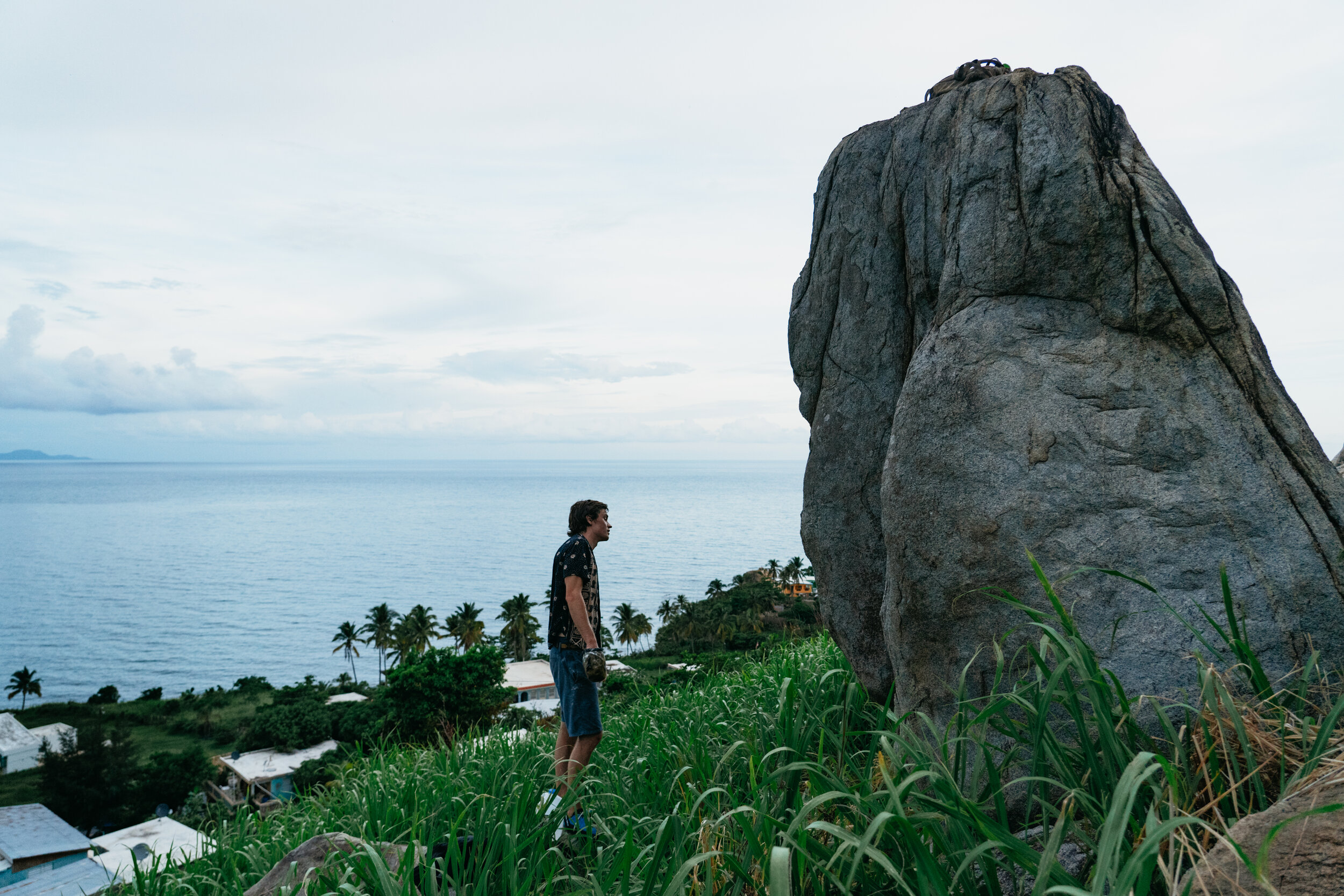 A man standing on a grassy hill overlooking the ocean with palm trees and houses in the background, facing a large rock formation.