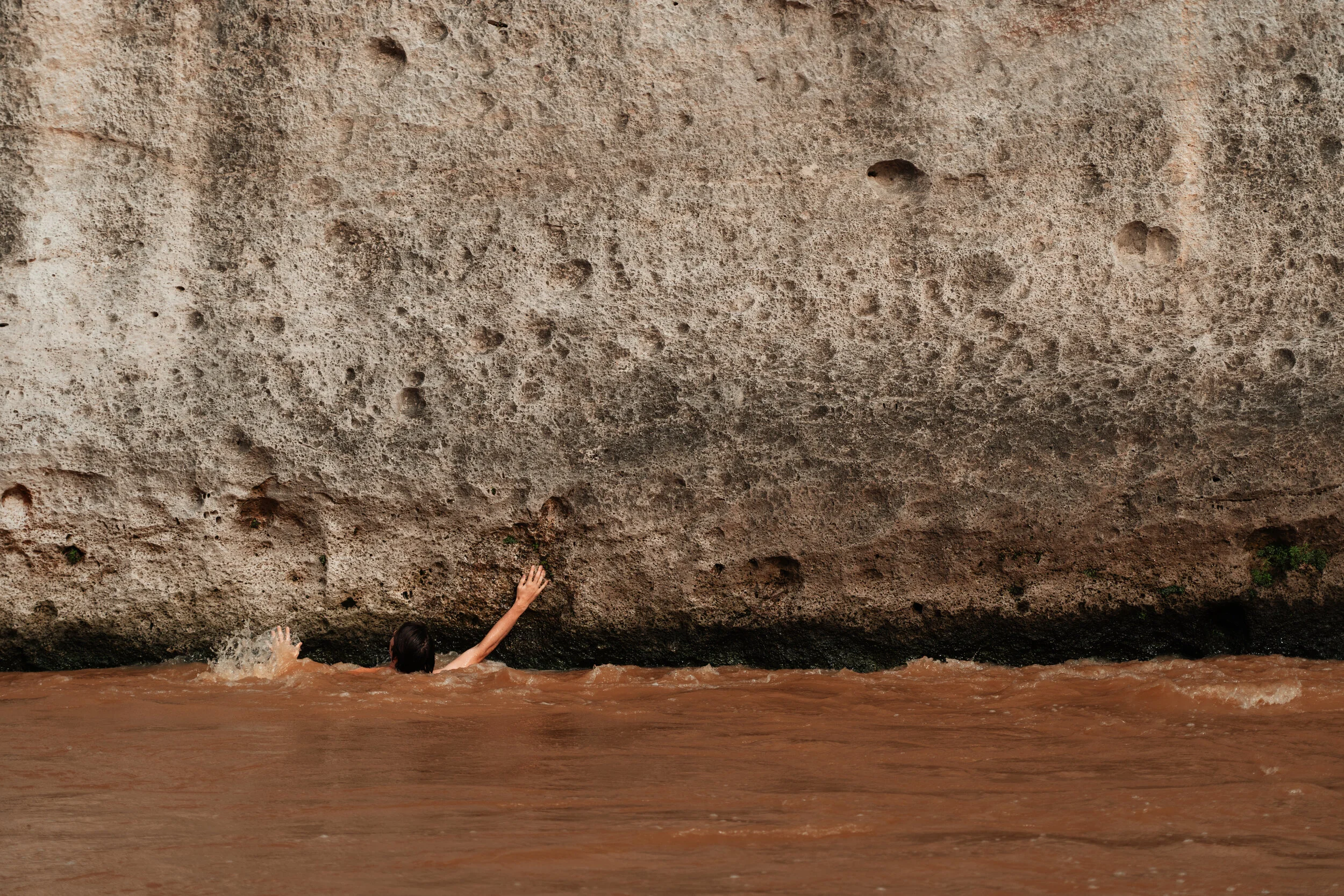 A person swimming in muddy water near a large, textured rock or cliff.