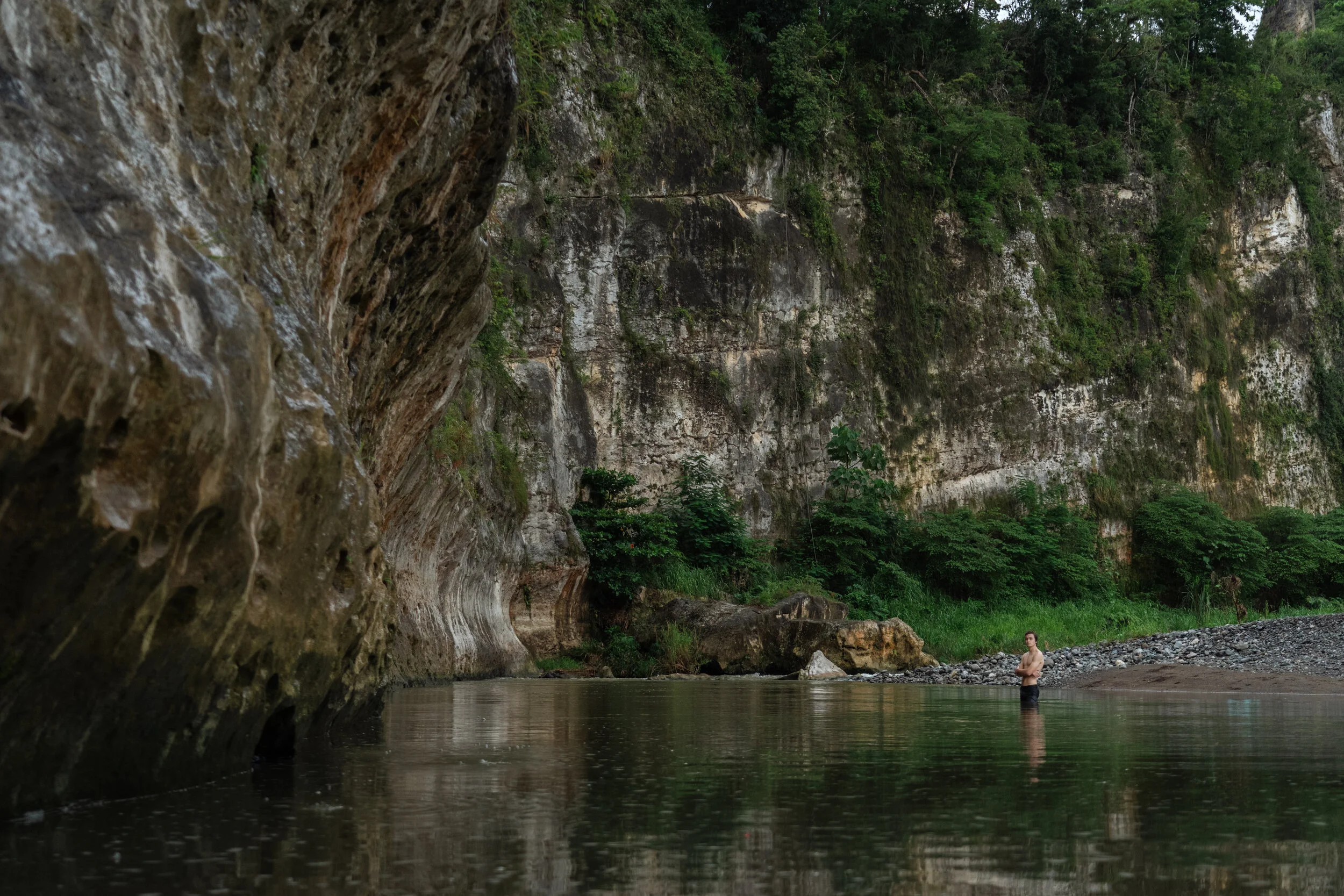 A man is standing in a body of water near a rocky cliff with lush green foliage in the background.