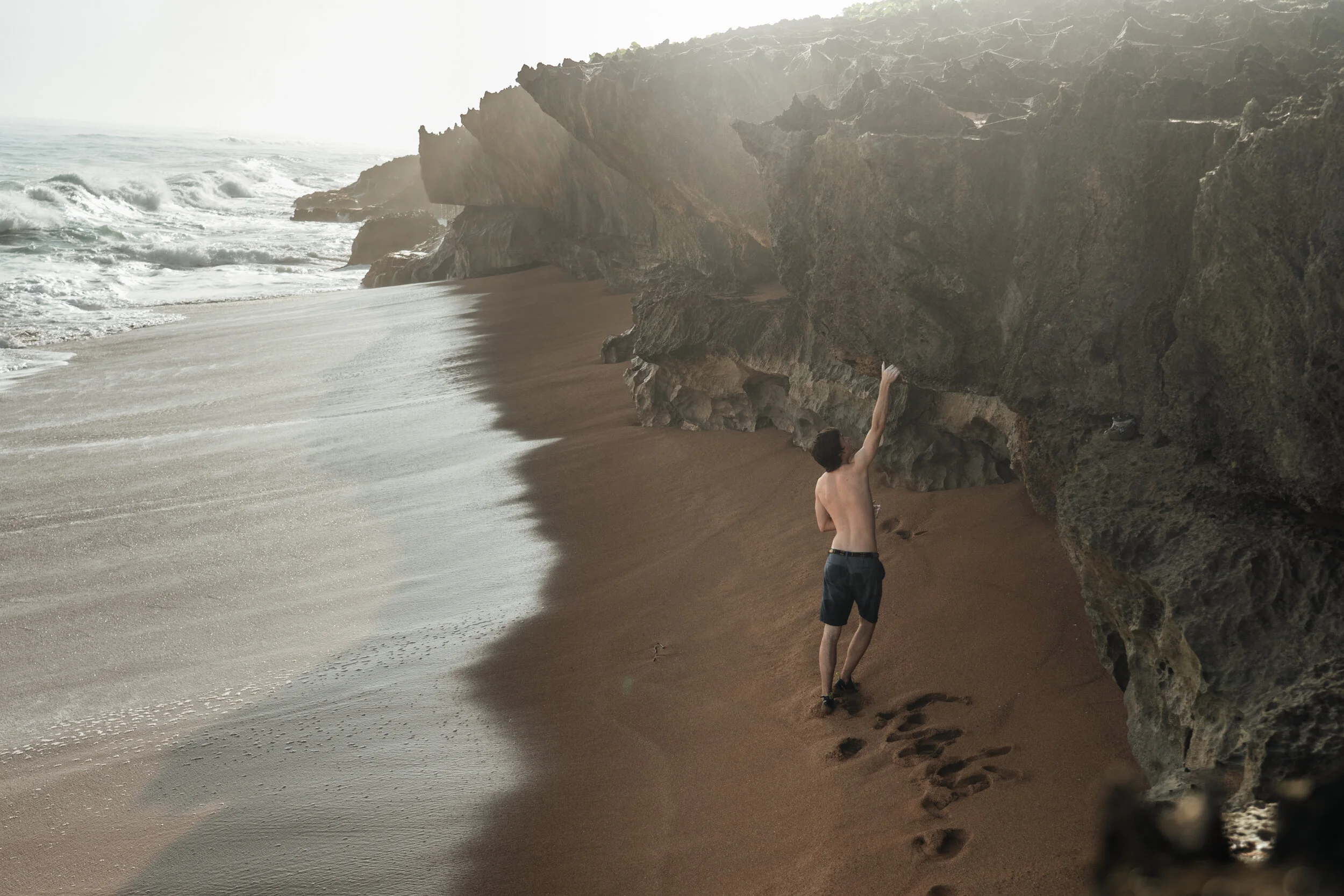 A shirtless man in black shorts walking along a sandy beach near large rocks, touching the rocks with one hand, with the ocean and waves in the background.