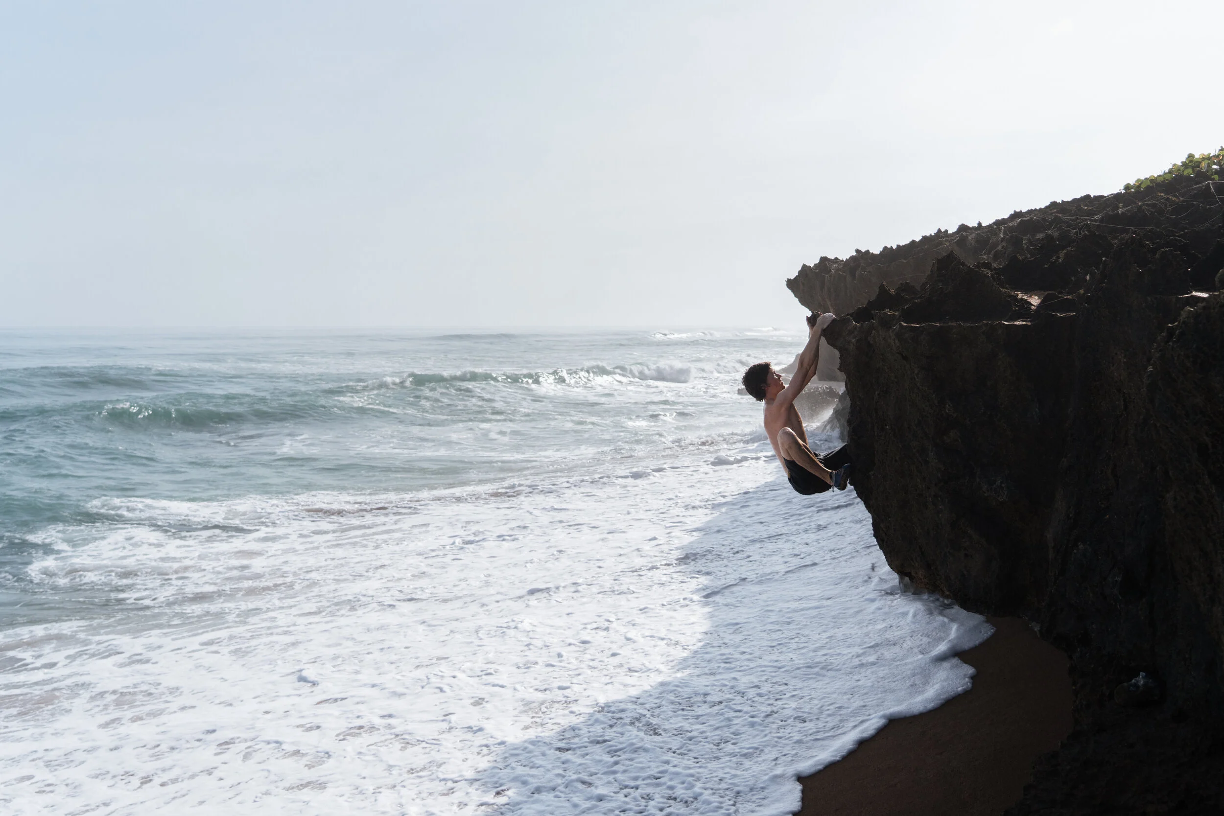 A person climbing a large rock by the ocean shoreline with waves crashing below.