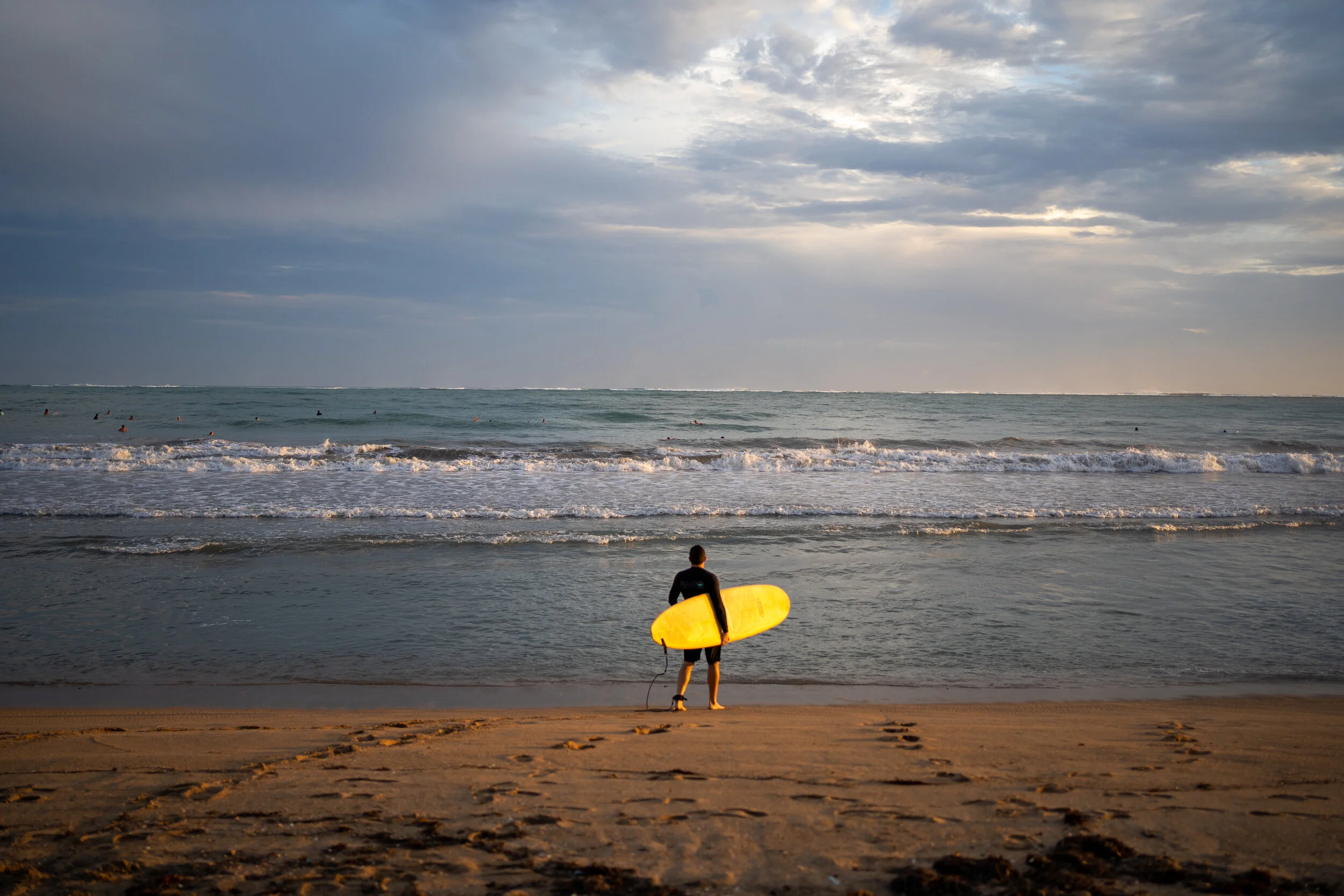 A person holding a yellow surfboard standing on the sandy beach facing the ocean, with several people surfing in the water under a cloudy sky.