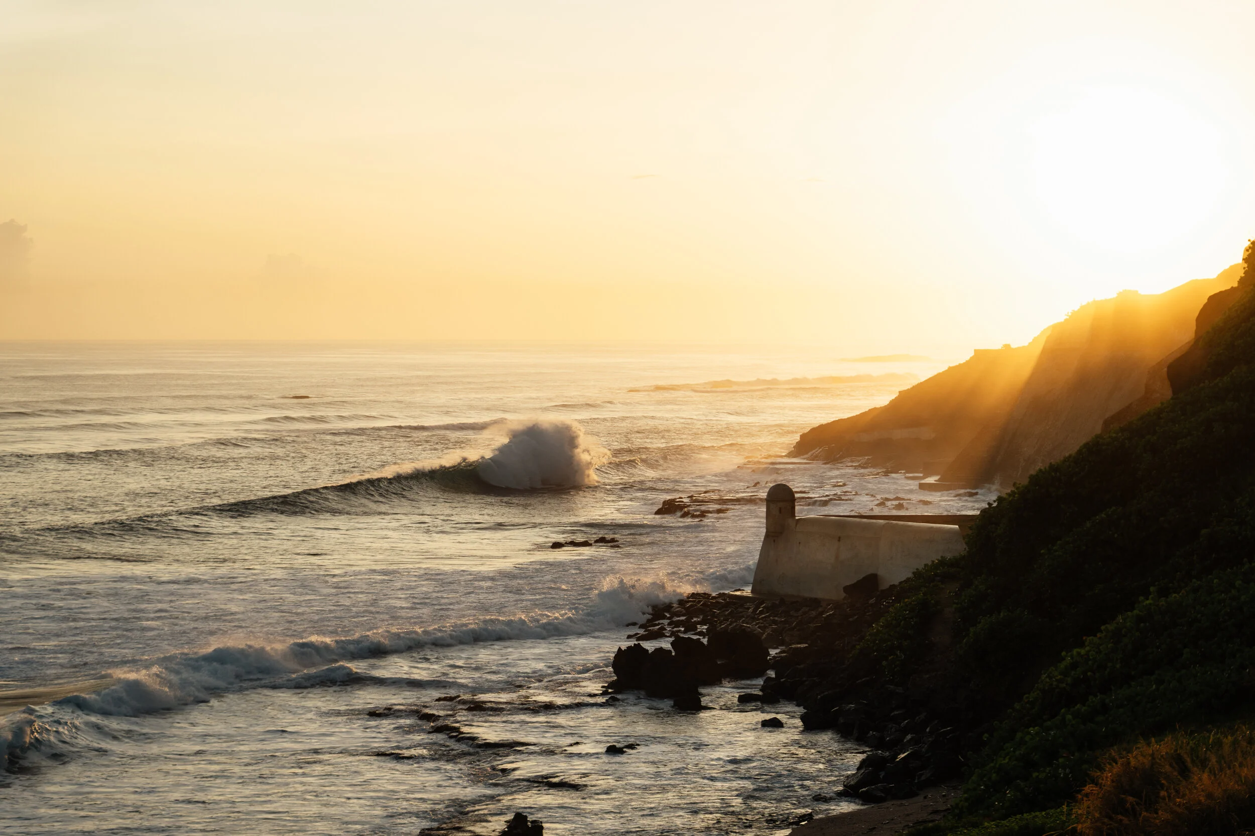Sunset over a rocky coastline with waves crashing, a lighthouse, and lush greenery.