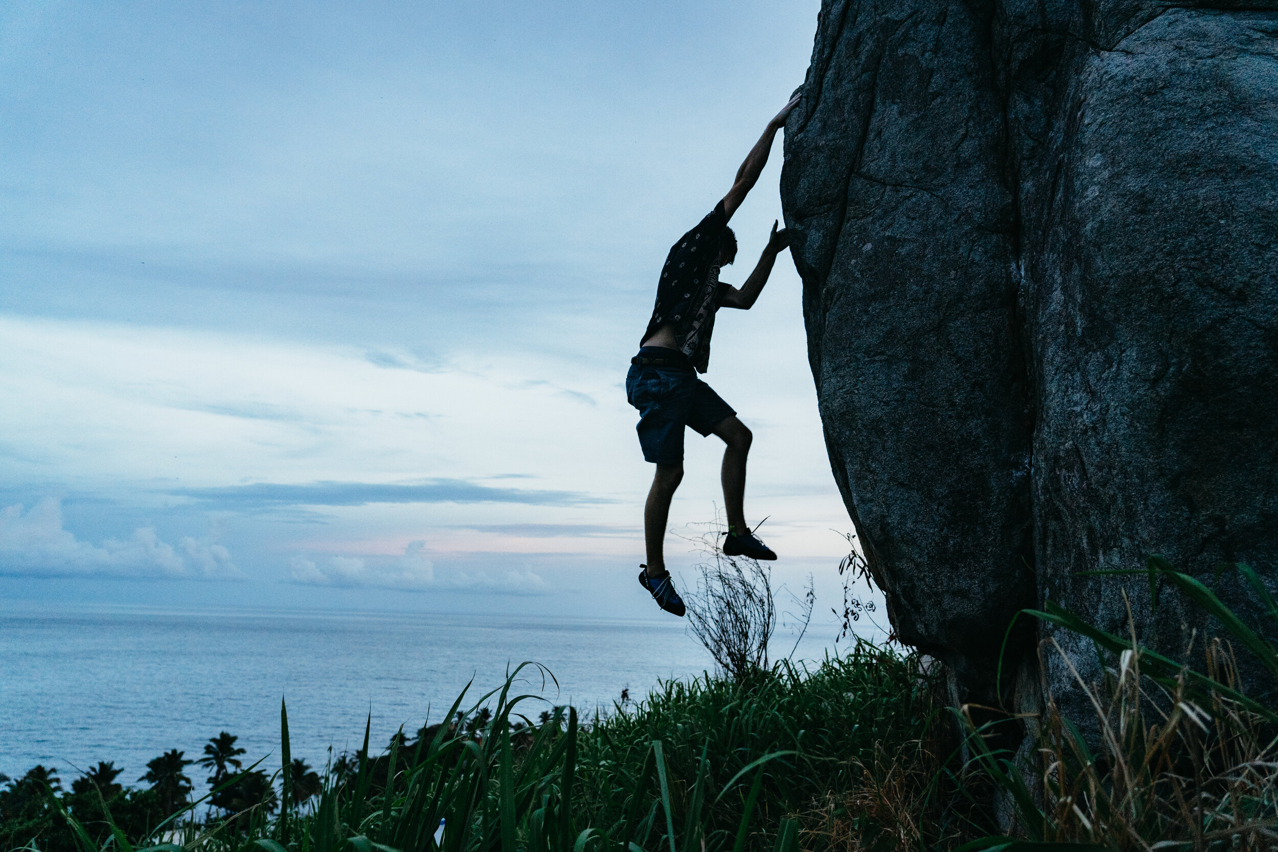 Person in black shirt and shorts climbing large rock near a body of water at dusk or dawn.