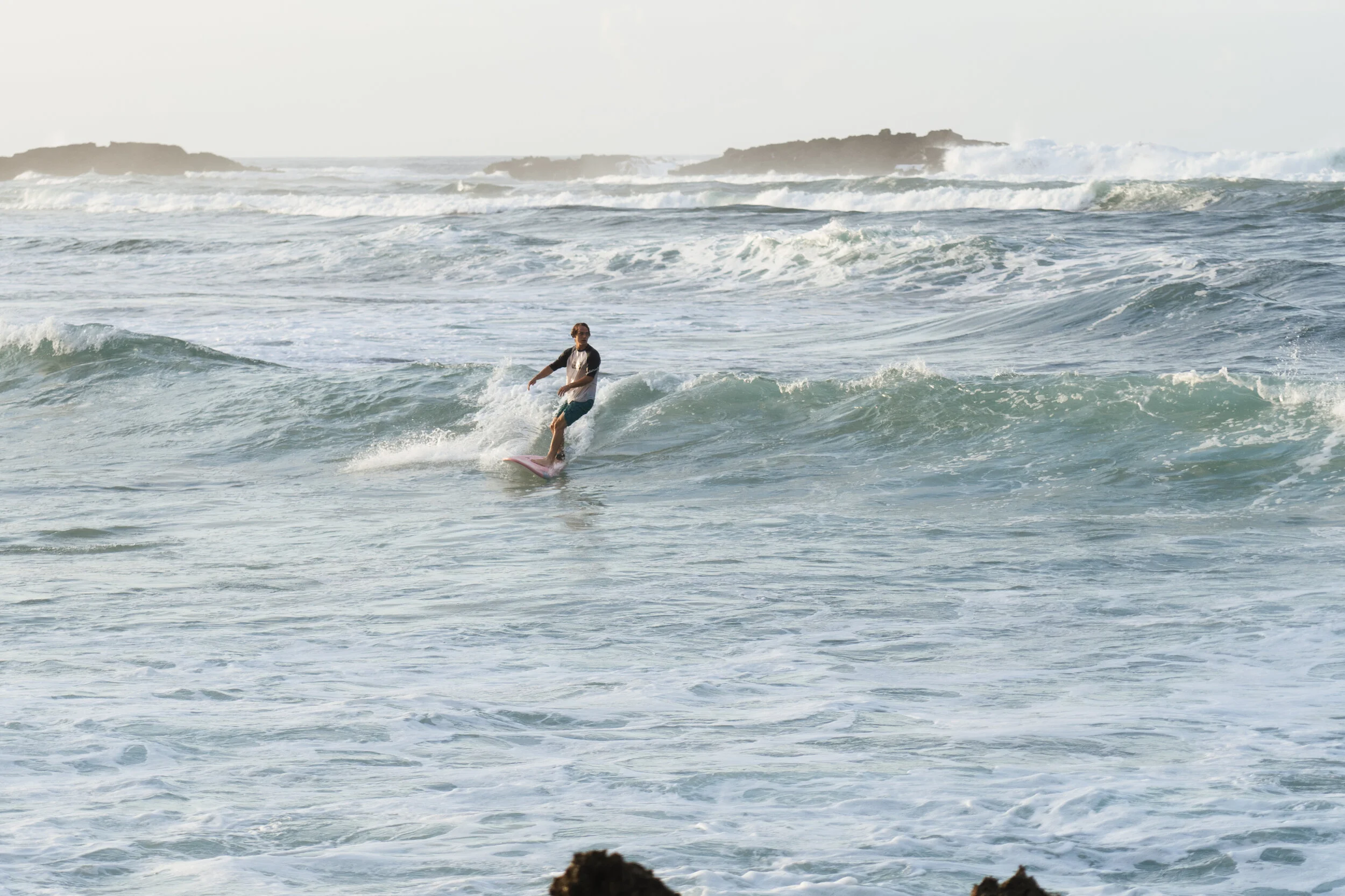 A person surfing on a pink surfboard in the ocean with waves and distant islands in the background