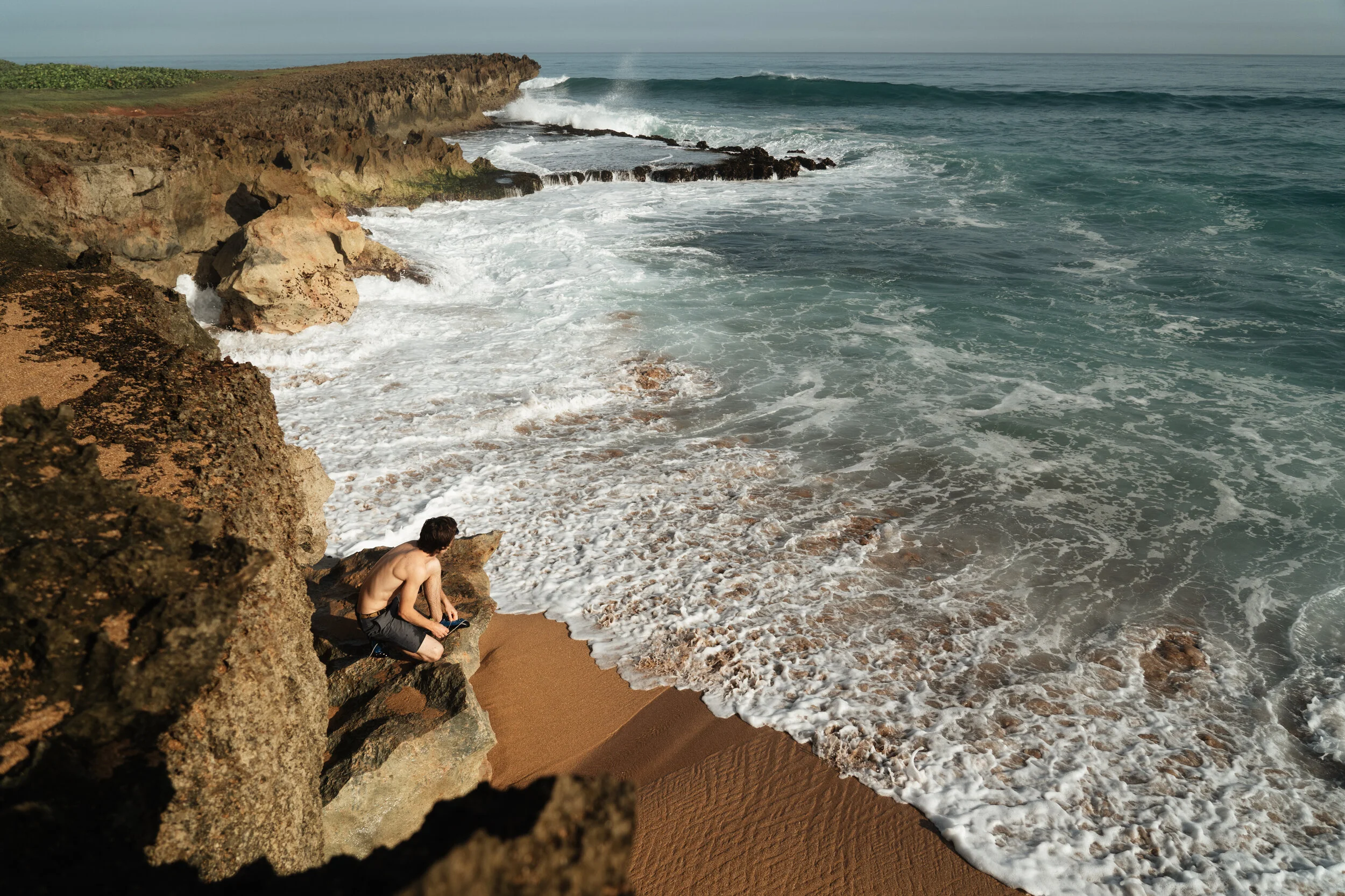 A shirtless man sitting on rocks at a beach with sandy shore and ocean waves crashing against the rocks.