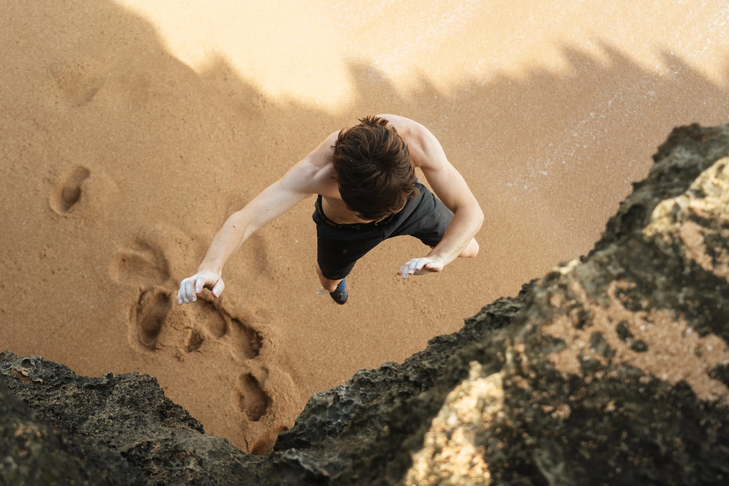 A shirtless man climbing a rocky cliff on a sandy beach, with footprints visible in the sand below.