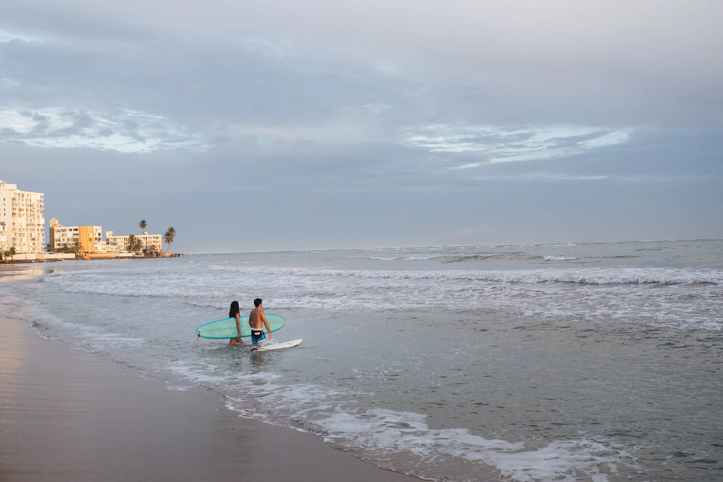 A couple walking into the ocean with surfboards near downtown buildings and palm trees on a cloudy beach.