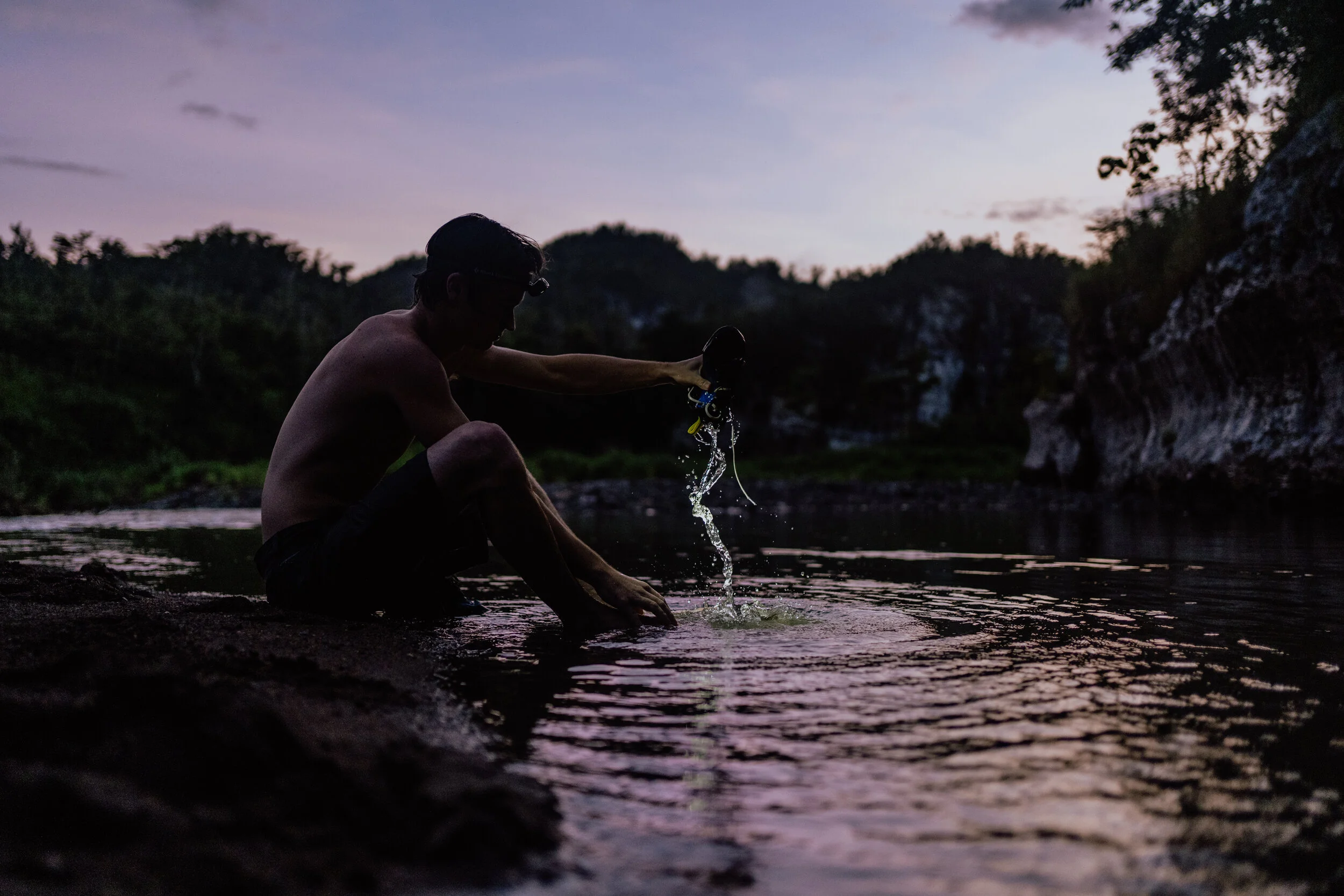 Person sitting by a river at dusk, pouring water from a container into the river, surrounded by nature.