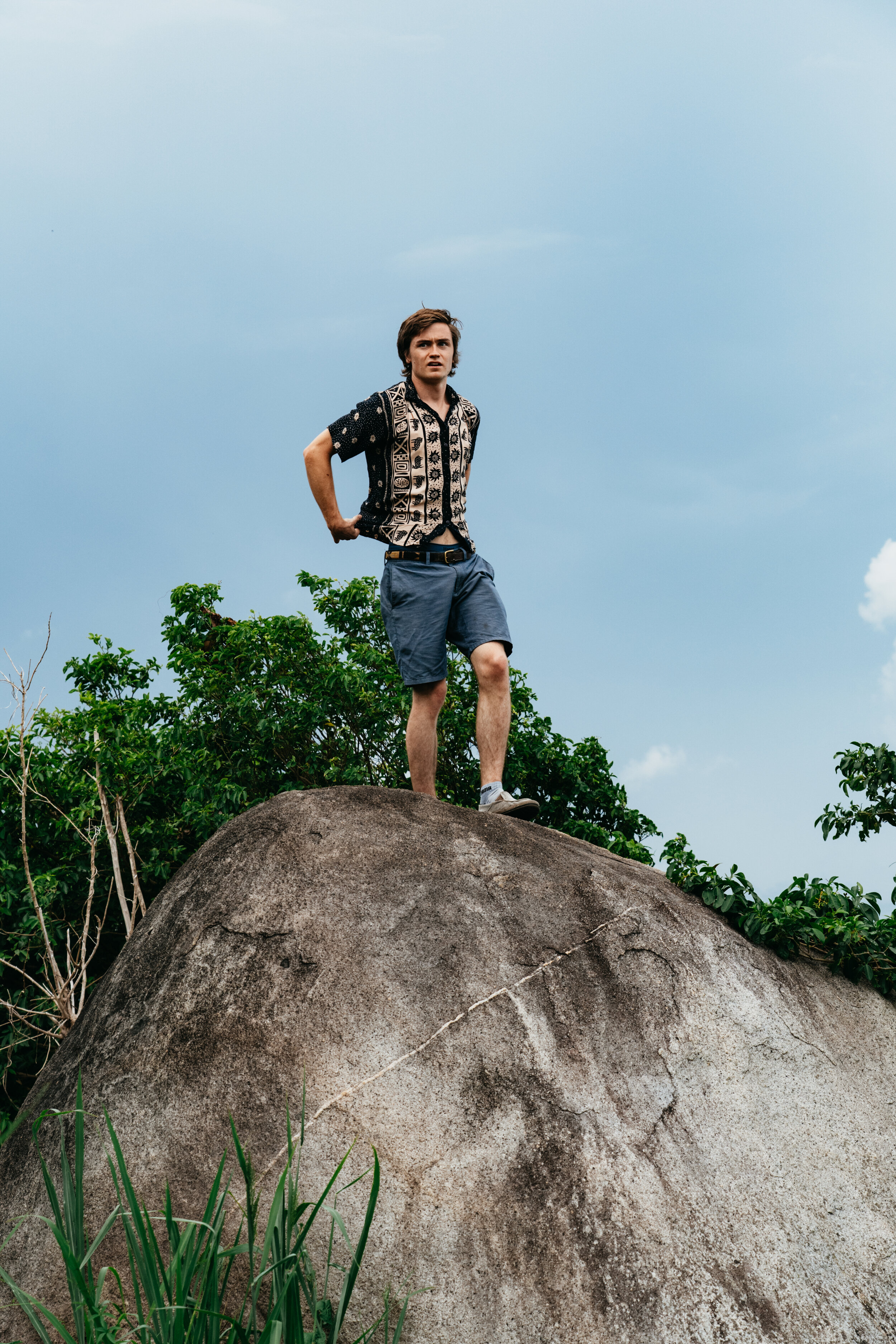 A young man standing on a large rock outdoors, with trees and a cloudy sky in the background.