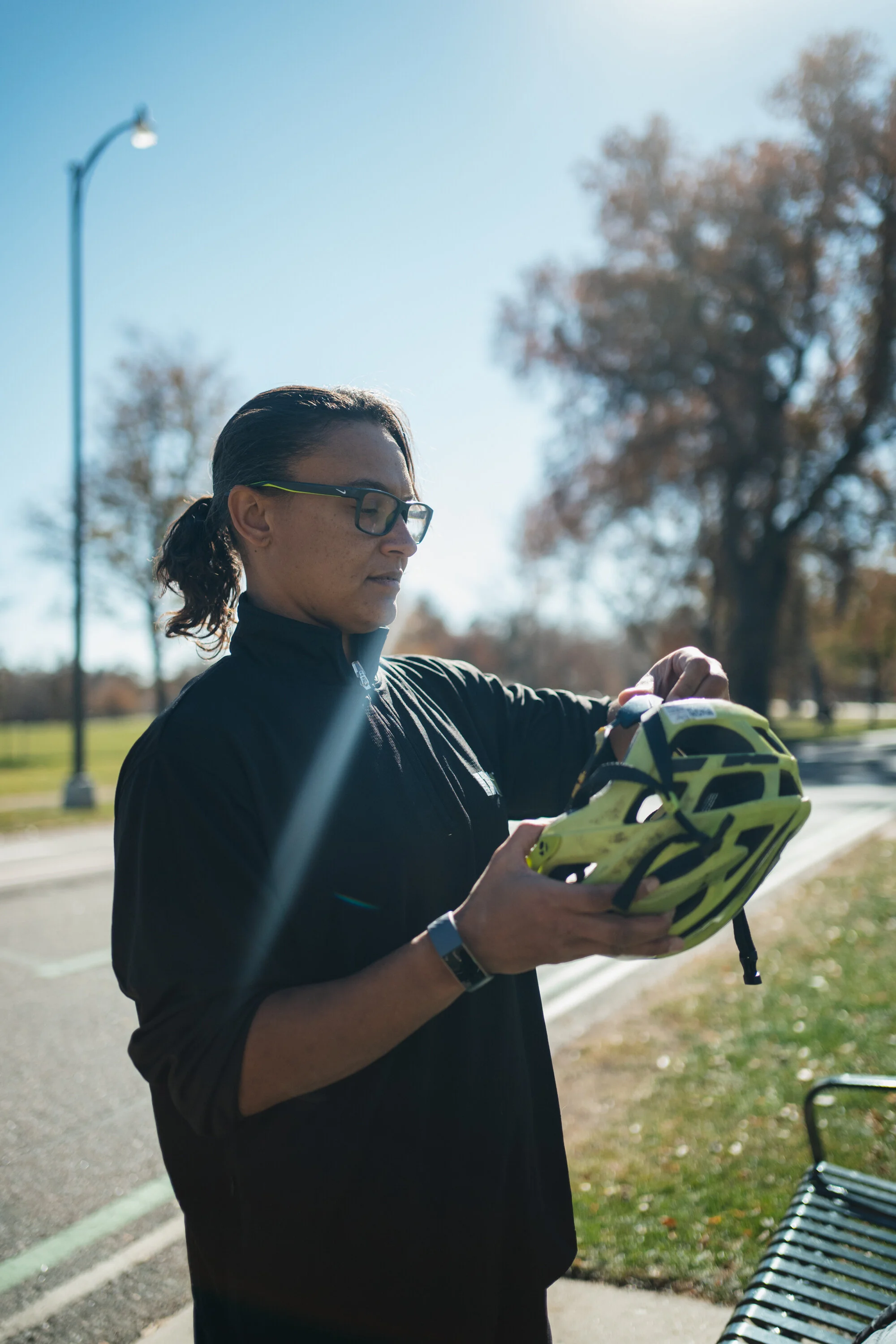A woman in black athletic clothing and glasses holding a yellow and black bicycle helmet outdoors on a sunny day