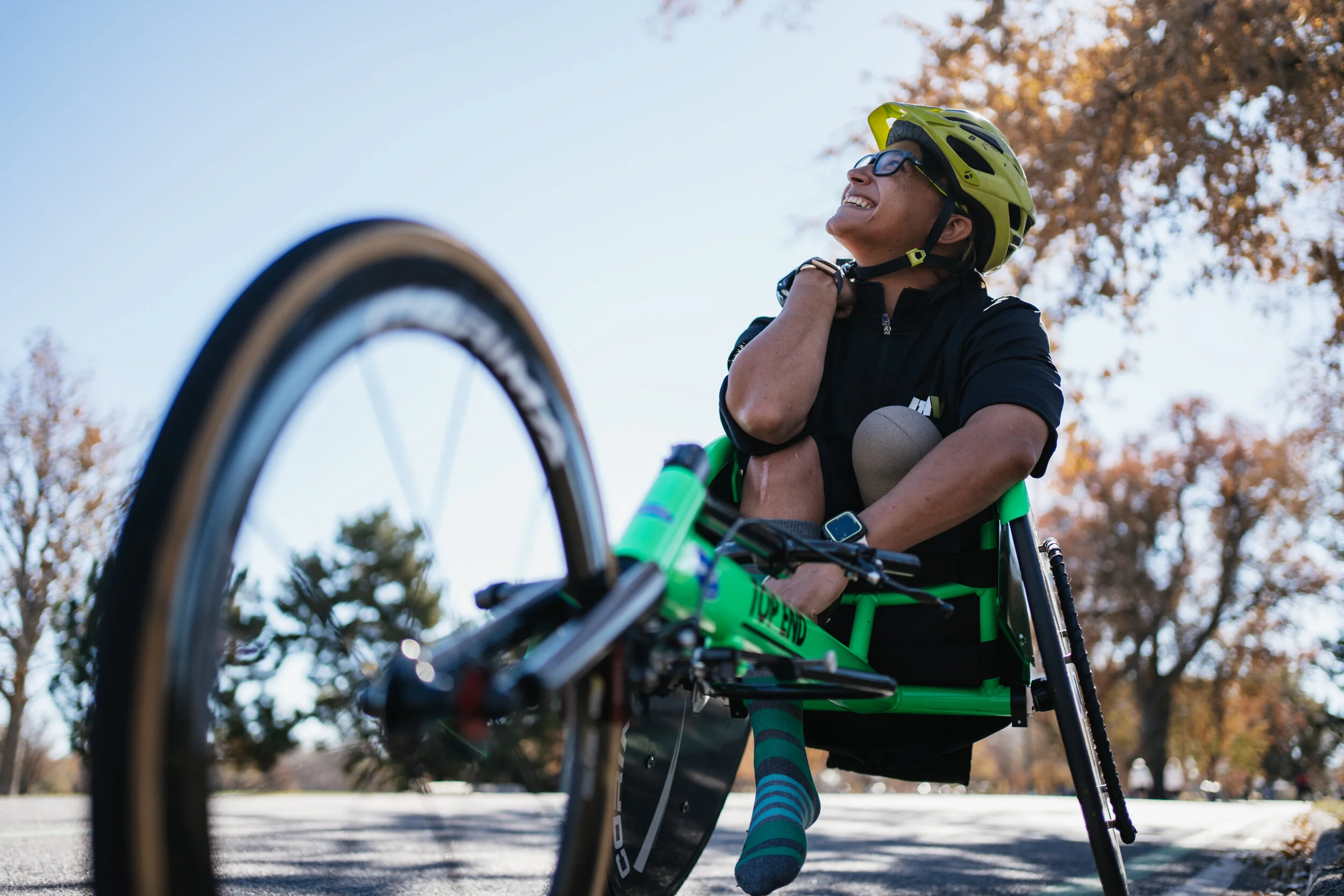 A woman in a wheelchair smiles while outdoors wearing a helmet, glasses, black shirt, and colorful socks, with trees and blue sky in the background.