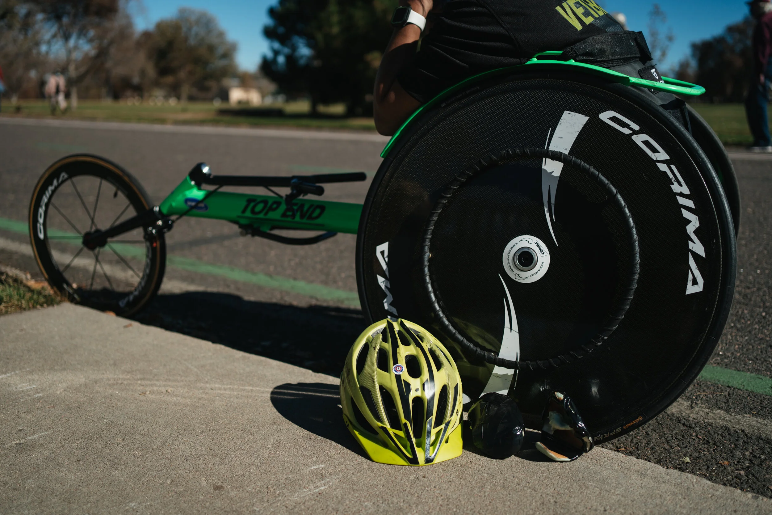 A green recumbent bike with a large black rear wheel resting on the sidewalk. A yellow bike helmet and black sunglasses are placed on the ground near the bike. The background shows a park with trees and people, under a sunny sky.