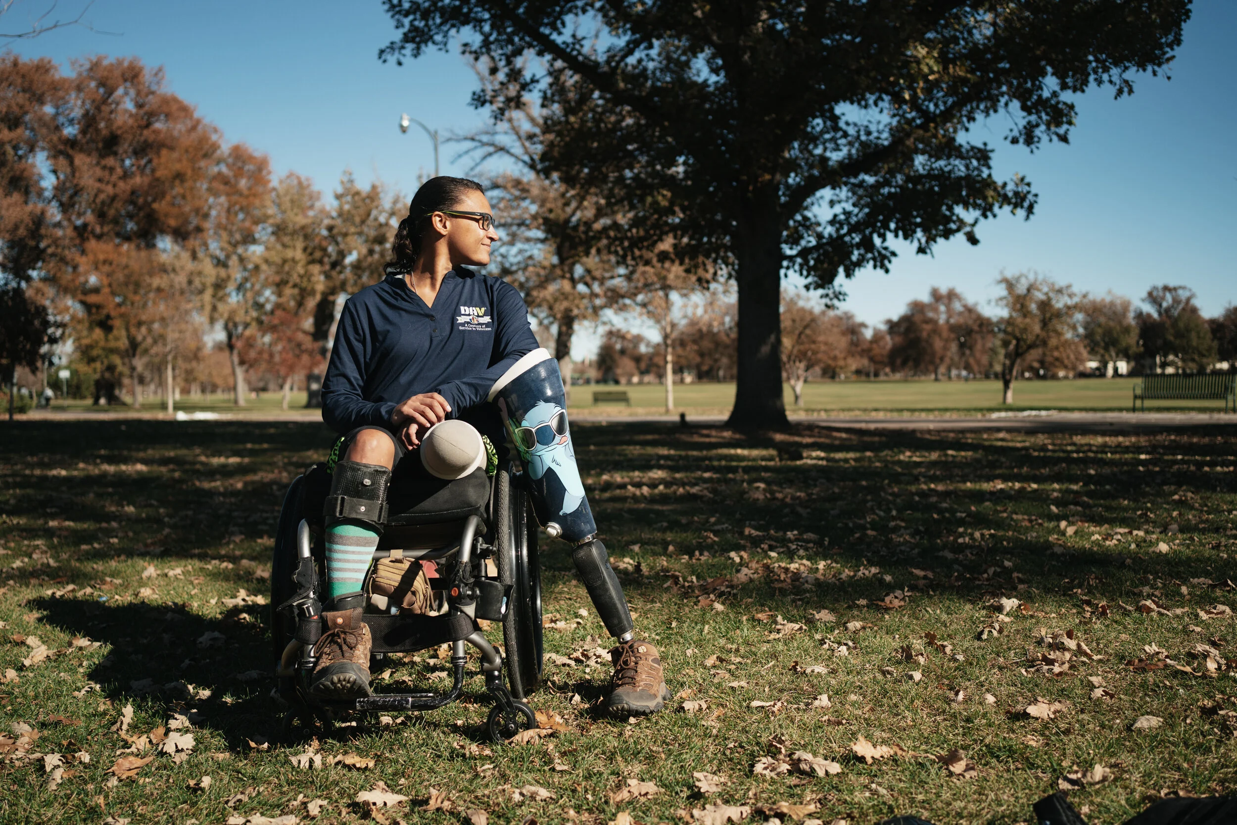 A woman in a wheelchair outdoors in a park, wearing a navy jacket and glasses, looking to the side with trees and a clear sky in the background.