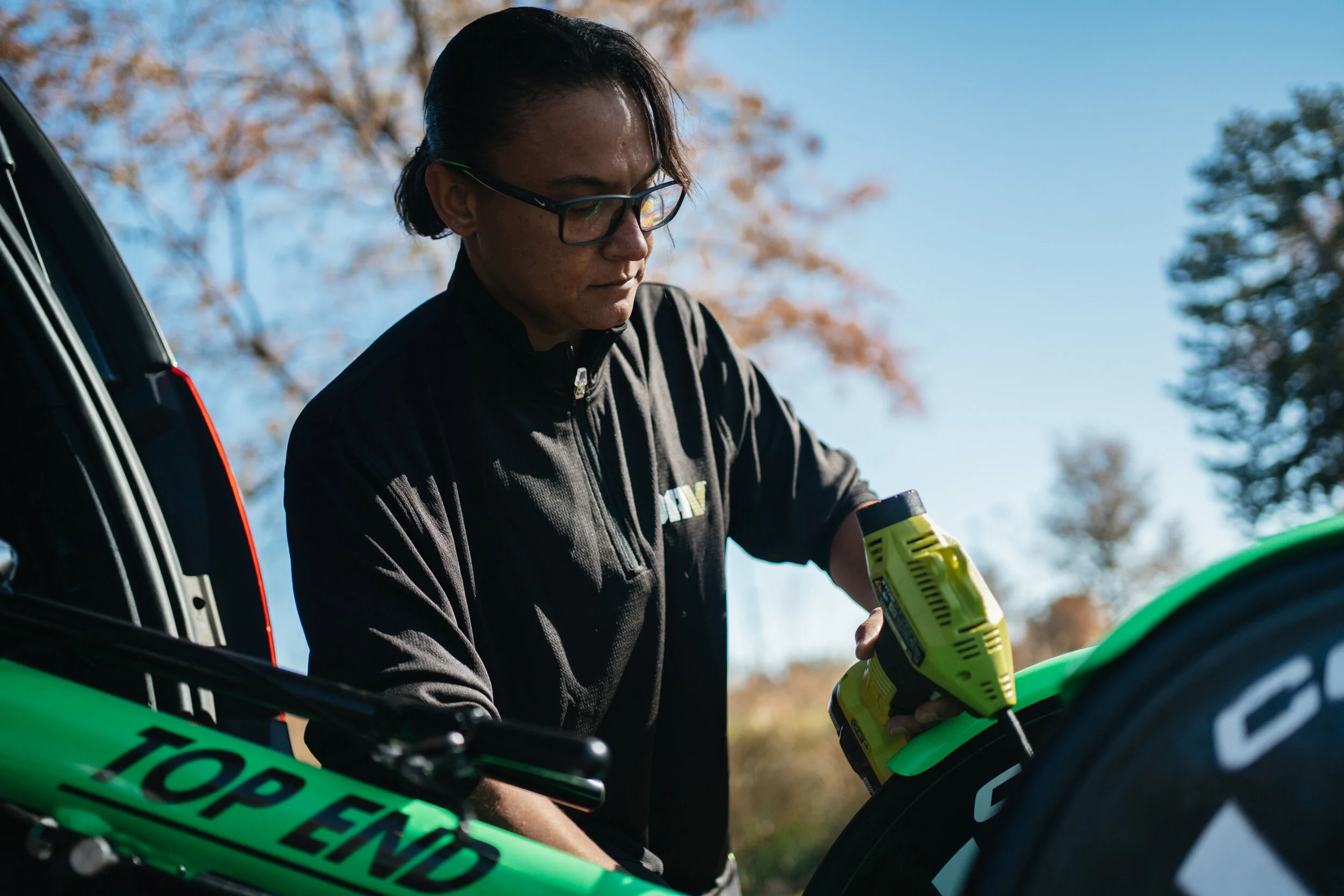 A person with glasses in a black jacket working on a green bicycle outdoors, using a yellow power tool.