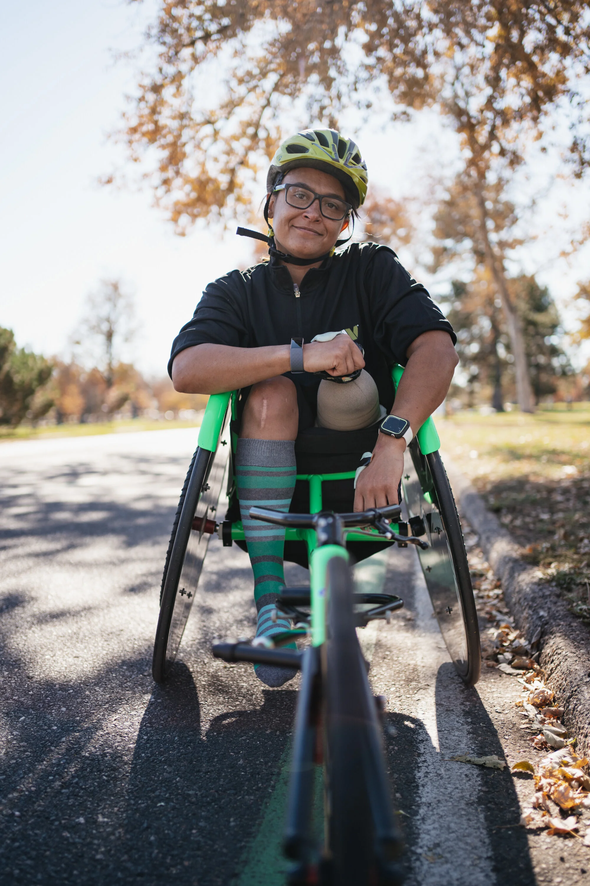 Person in a wheelchair wearing a helmet and glasses, sitting outside on a sunny day with trees in the background.