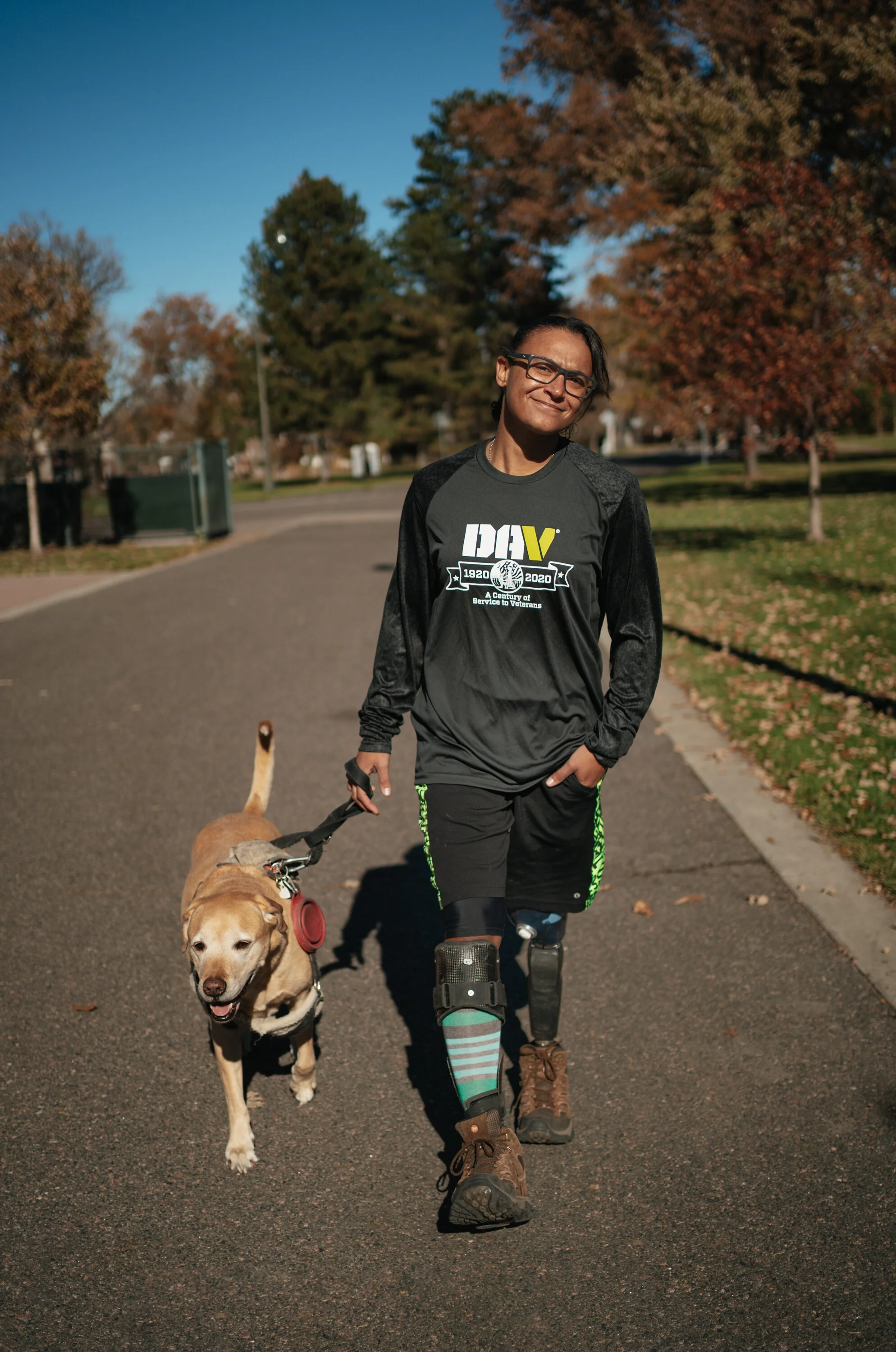 Person with prosthetic leg wearing outdoor clothing walking a service dog on a leash in a park during autumn.