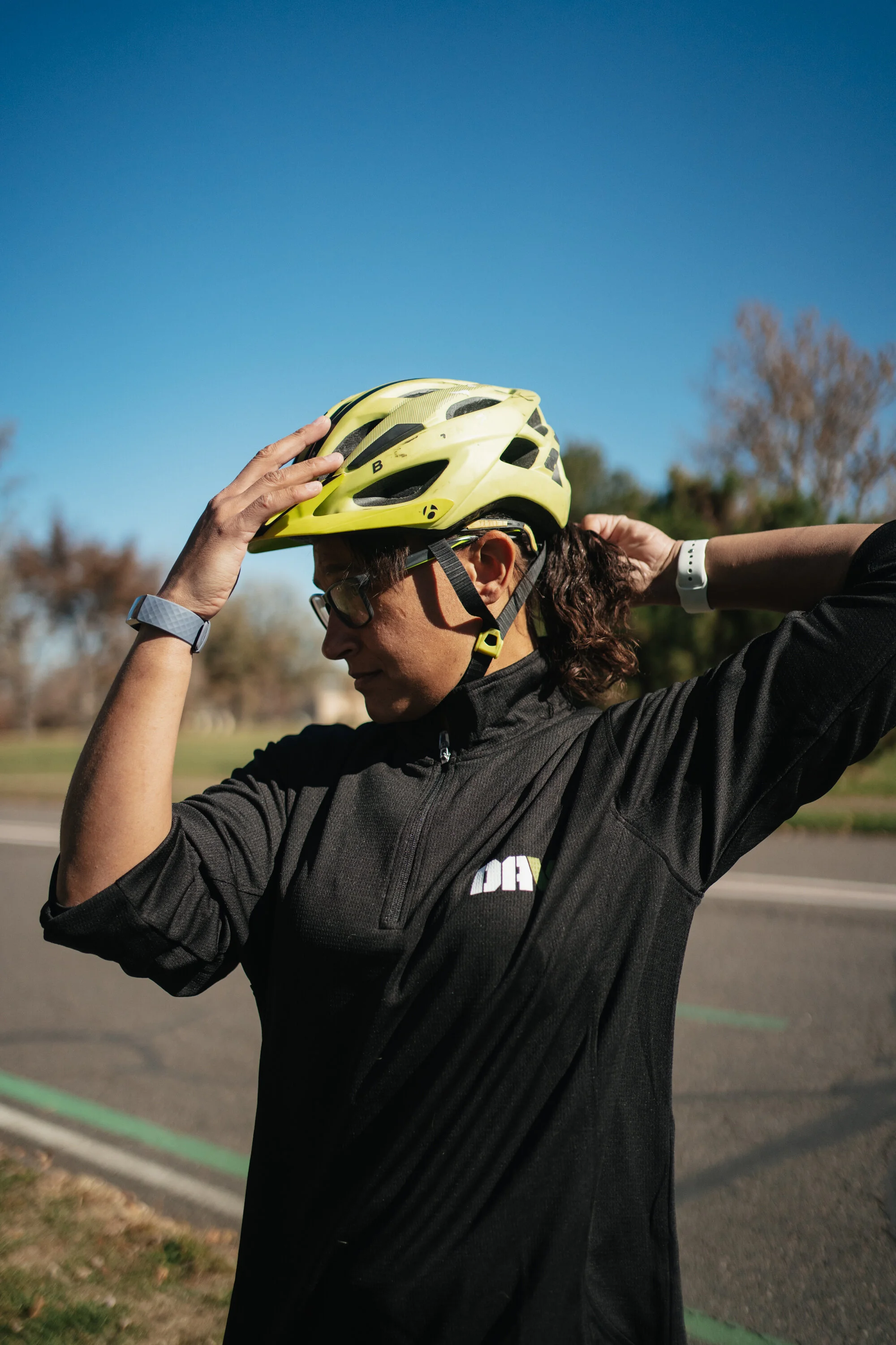 A woman adjusting her yellow bike helmet outdoors on a sunny day, standing on a paved surface with trees in the background.