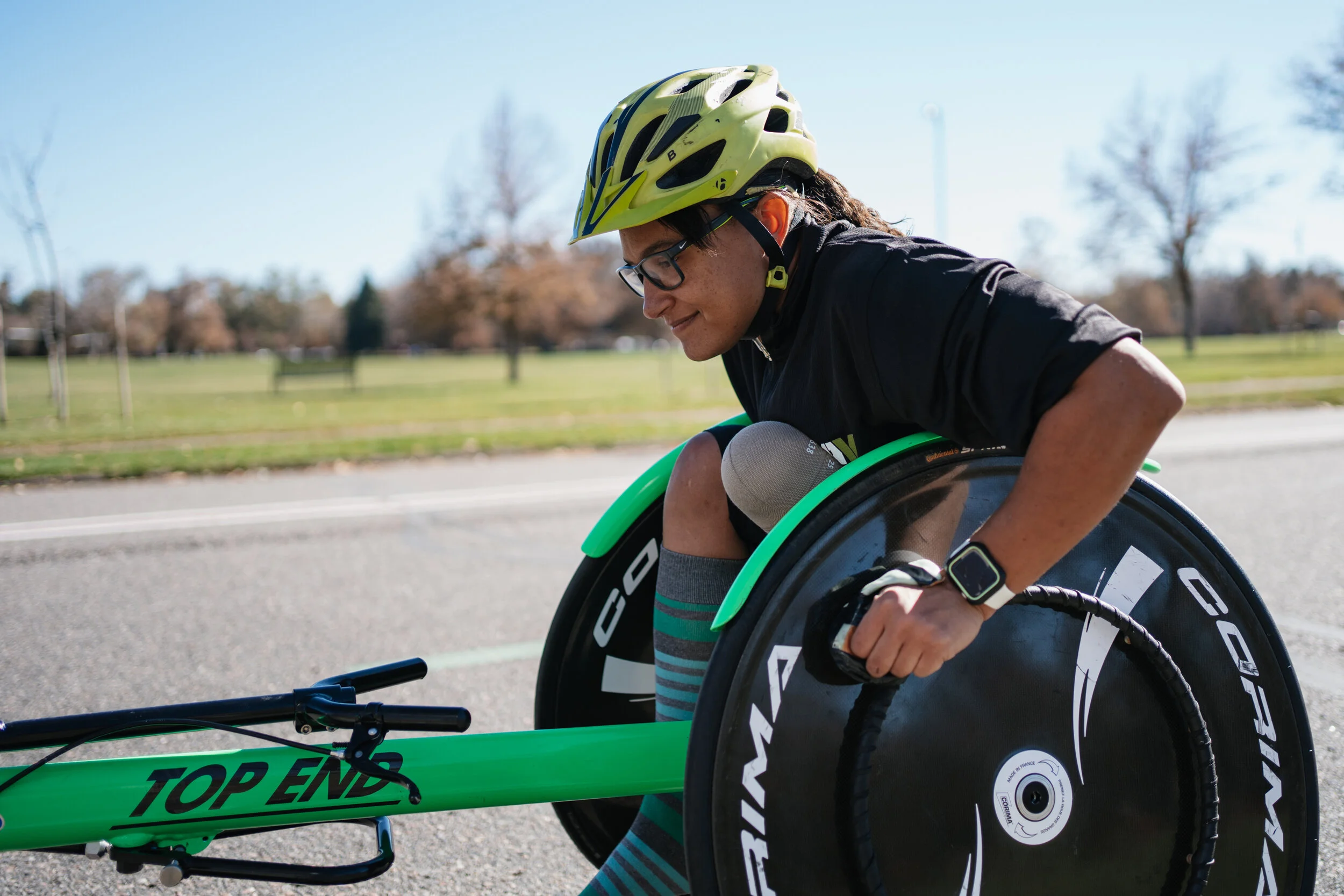 A person wearing a yellow helmet and glasses prepares their green racing handcycle on a paved road in a park with trees and grassy fields in the background.