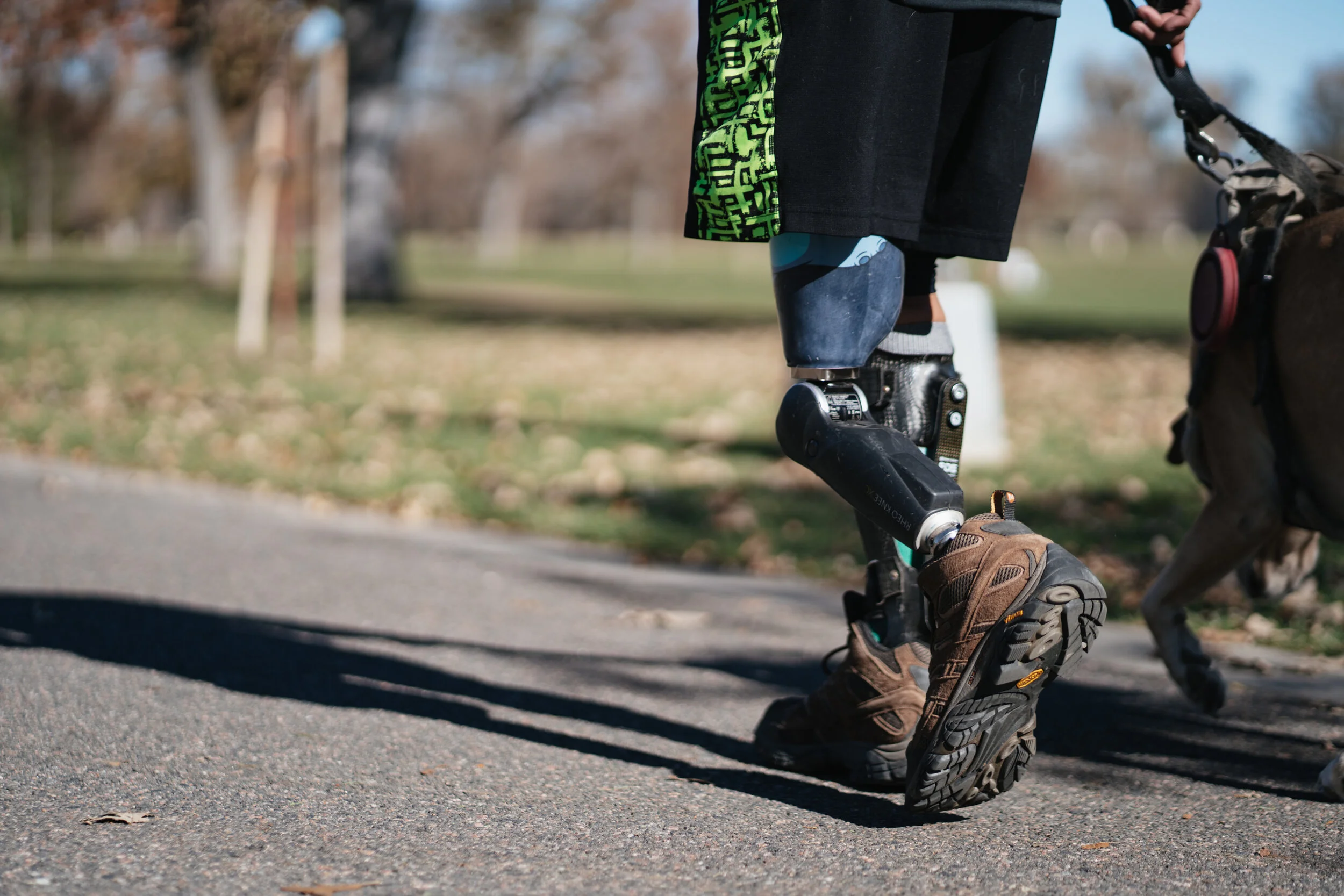 A person with a prosthetic leg walking a dog in a park on a paved path, with trees and fallen leaves in the background.