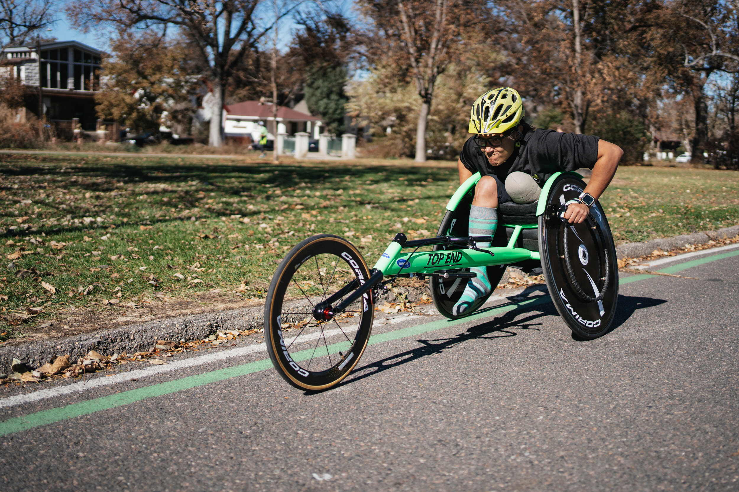 A person in a racing wheelchair with a green frame and black wheels, wearing a yellow helmet and glasses, leaning forward as they move quickly on a paved road in a park, with autumn trees and houses in the background.