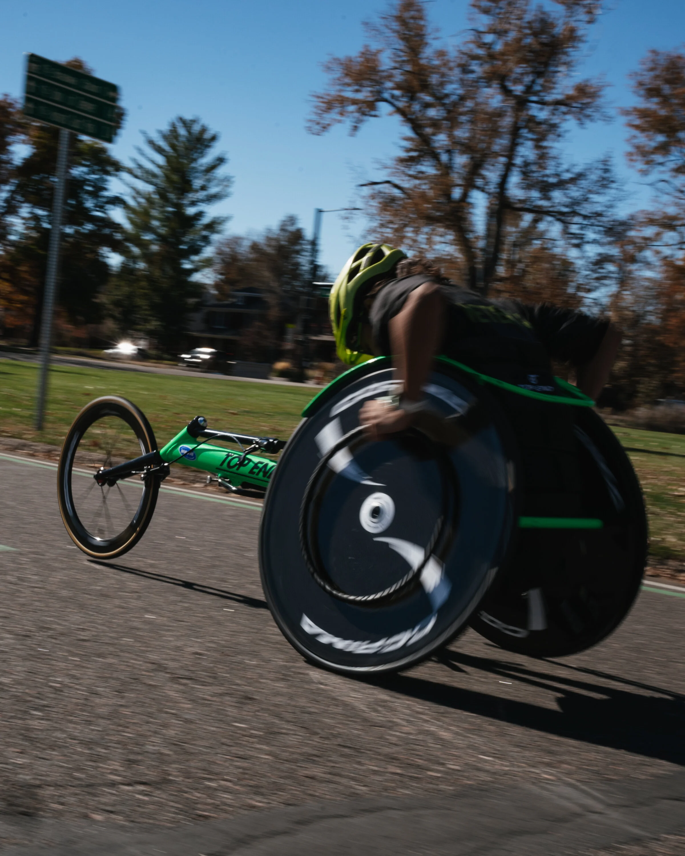 Person in a wheelchair, wearing a helmet, racing on a paved path with trees and a few cars in the background.