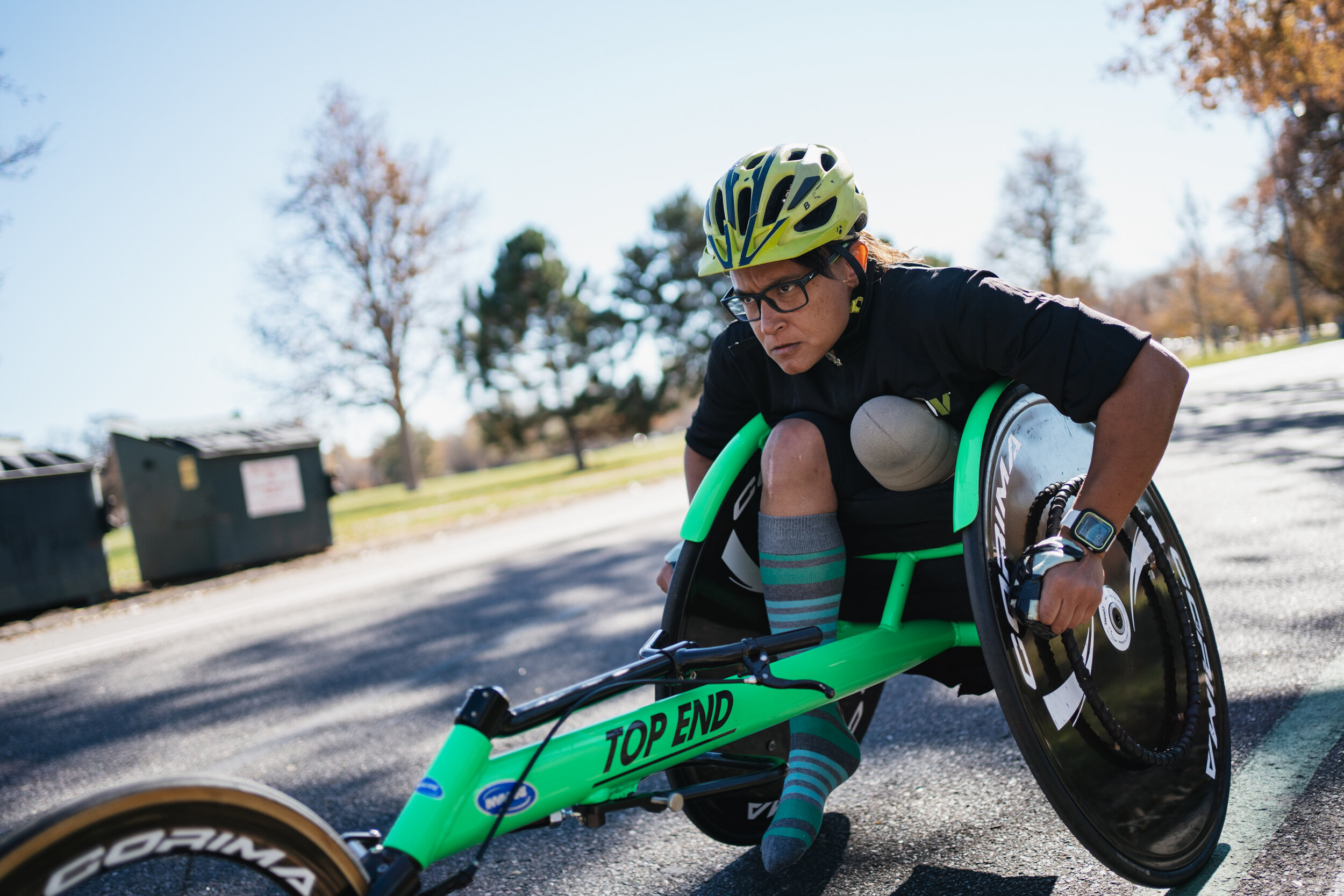 A man with glasses and a yellow helmet riding a green handcycle outdoors on a sunny day.