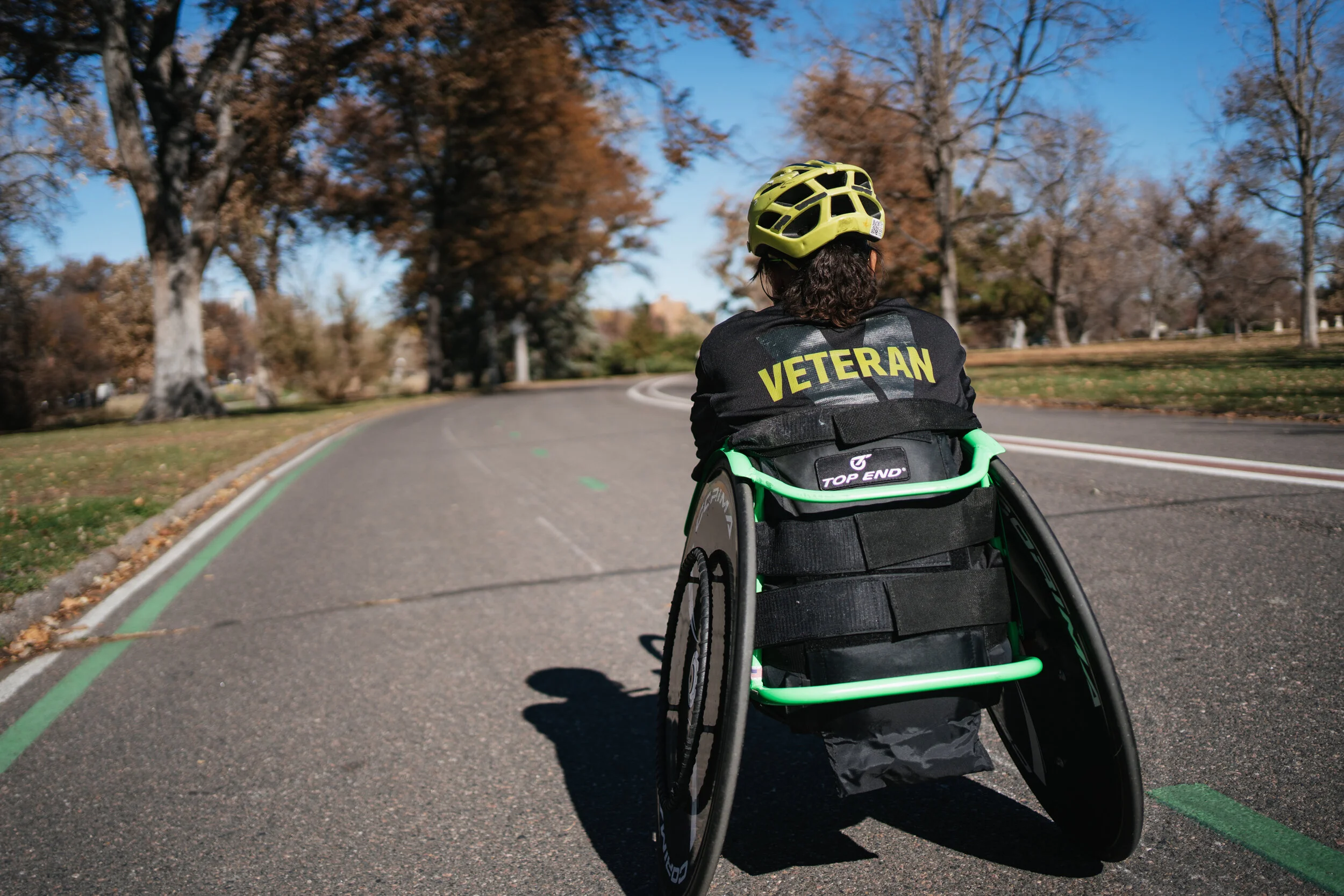 Person in a wheelchair riding on a paved outdoor path surrounded by trees with autumn leaves, wearing a yellow helmet and a black shirt with 'VETERAN' printed on the back.