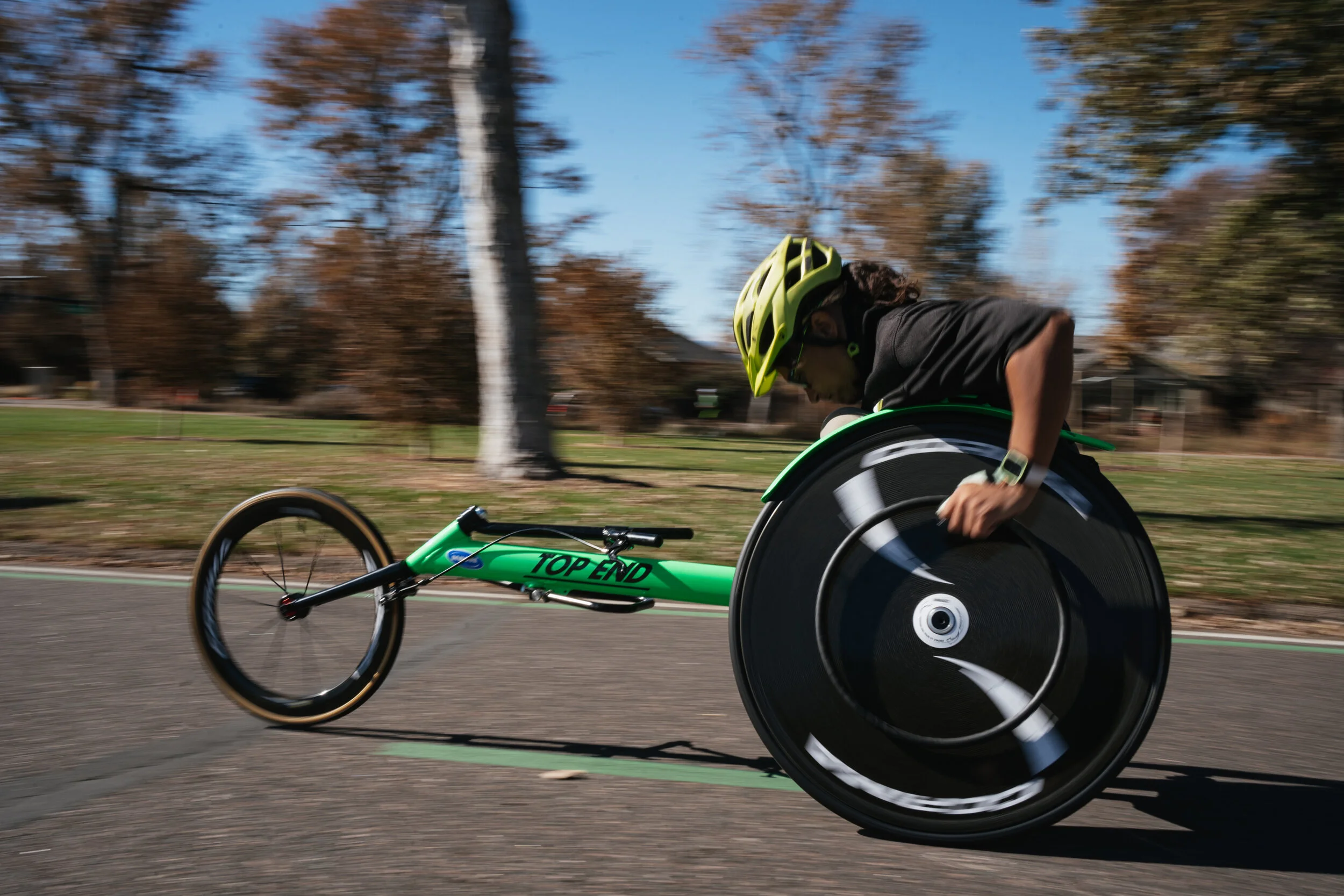 Person in a green helmet racing on a green adaptive bike with large black wheel on a paved path in a park with autumn trees.