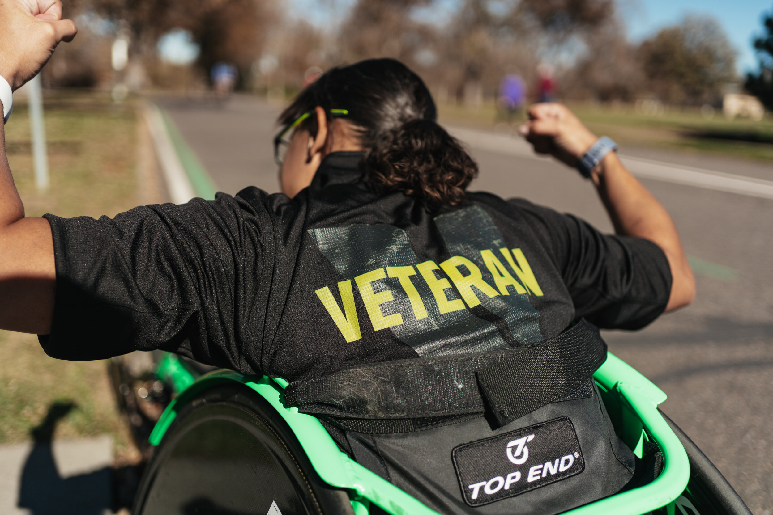 A person with dark hair tied back, wearing sunglasses and a black jacket, is viewed from behind in a wheelchair with a green frame. The jacket has the word 'VETERAN' on the back. The person is flexing their arms in a pose of strength outdoors on a su