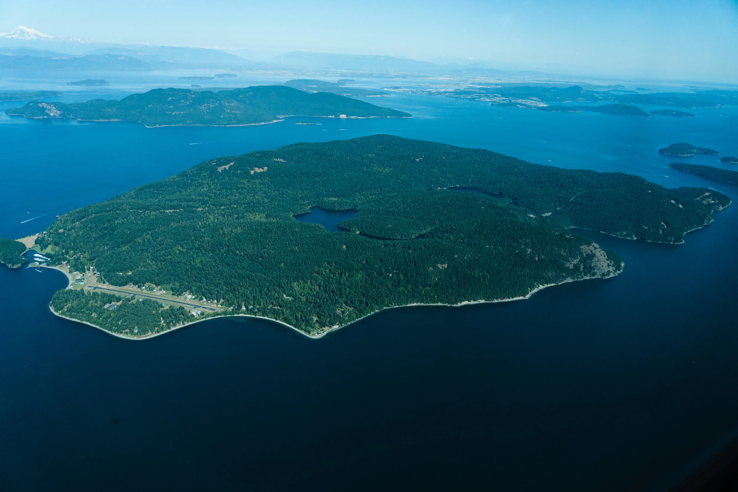 Aerial view of a large, forested island surrounded by a body of water, with smaller islands in the distance, and mountains in the background.