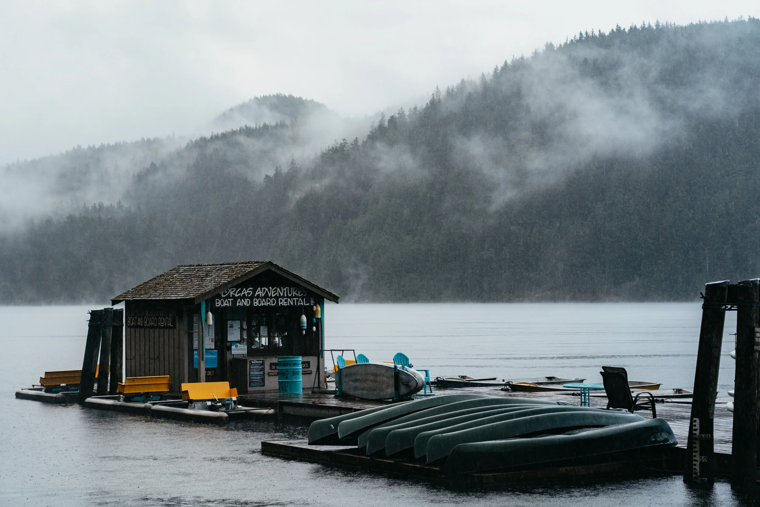 A floating dock with canoes, kayaks, and a small building that reads 'Oregon Adventures Boat and Board Rental' on a foggy lake surrounded by forested mountains.