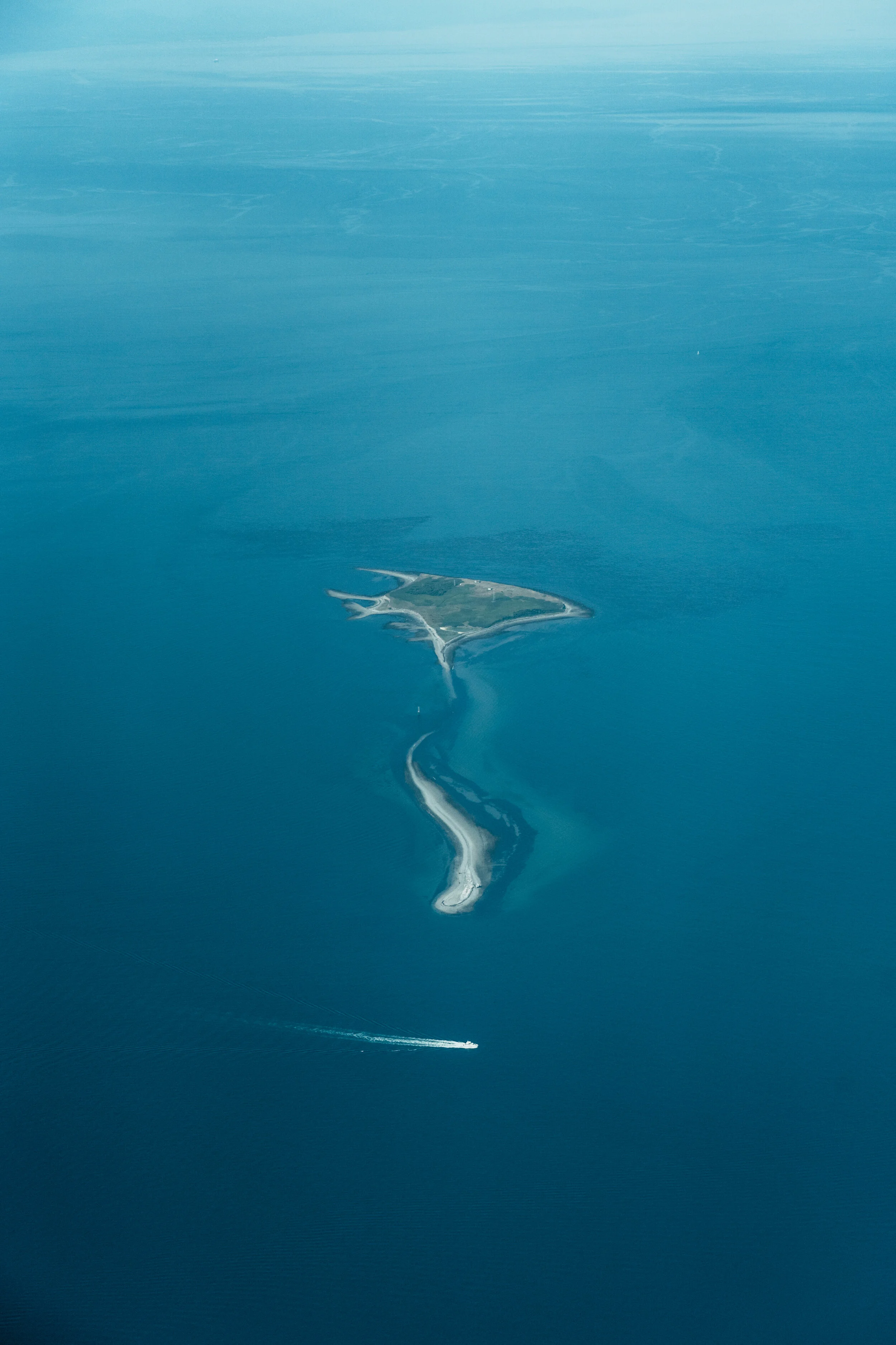 Aerial view of a narrow, winding island in the ocean with sandy shores and green land, and a boat leaving a wake in the water nearby.