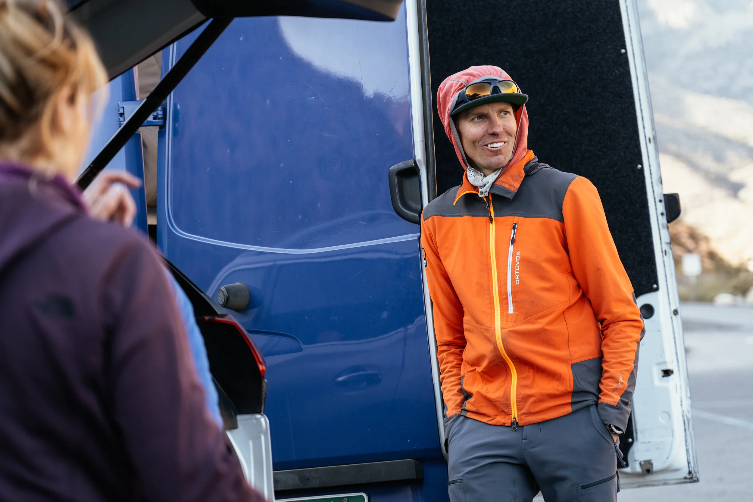 A man in outdoor gear smiling and talking to a woman near a blue van, with mountains in the background.